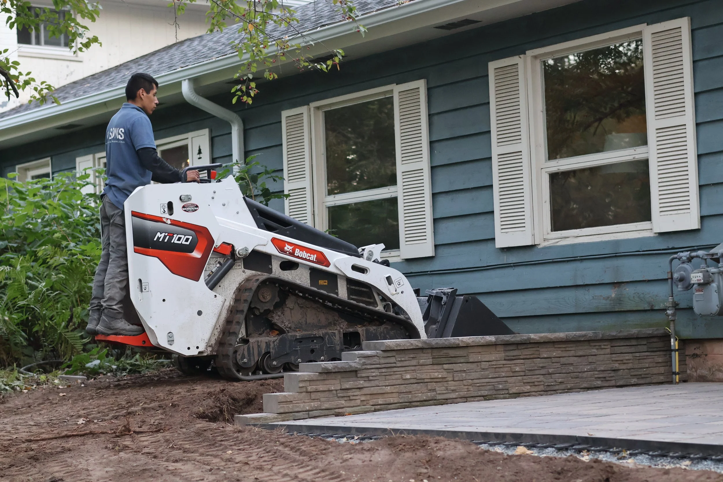 A worker operating a compact track loader in front of a house with blue siding and white window shutters, laying bricks or pavers for a sidewalk or patio area.