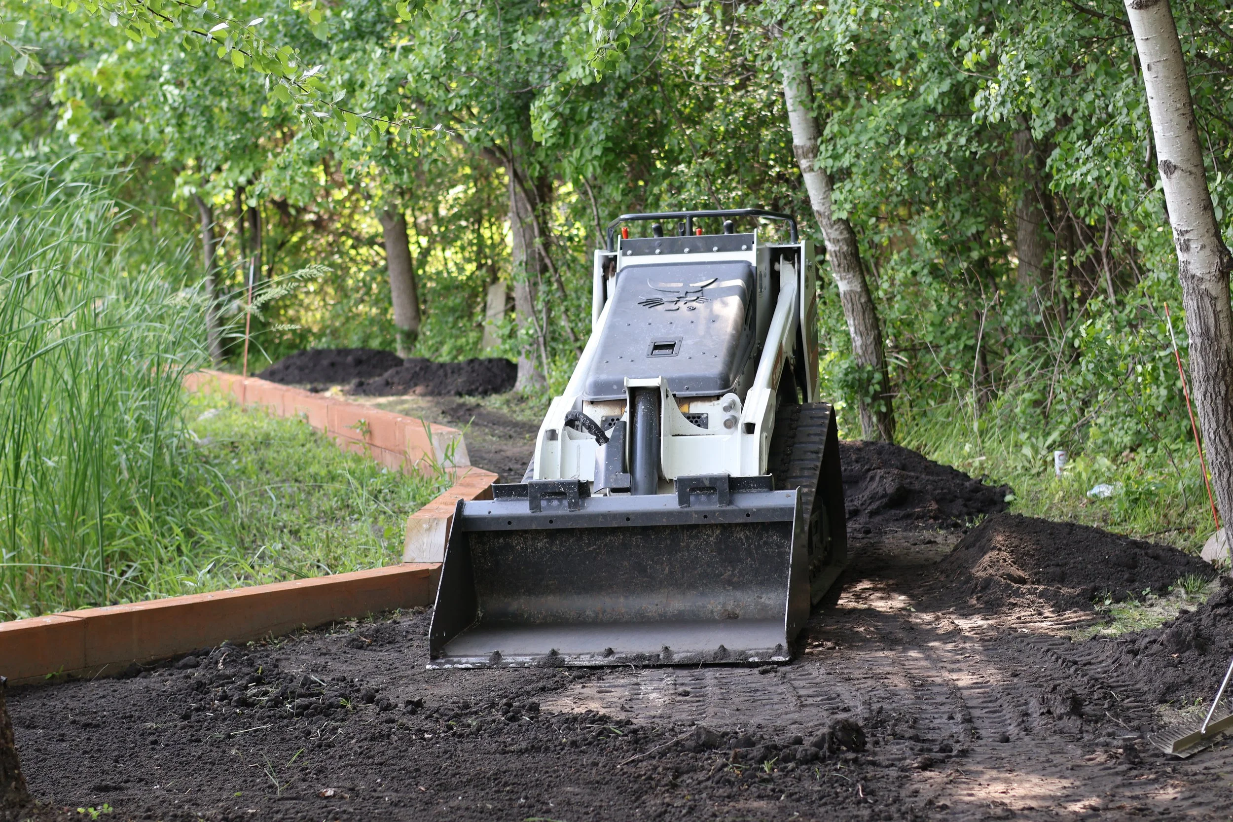 A small construction vehicle, possibly a mini loader or skid-steer loader, spreading soil or dirt along a landscaped garden bed lined with bricks, surrounded by green trees and grass.