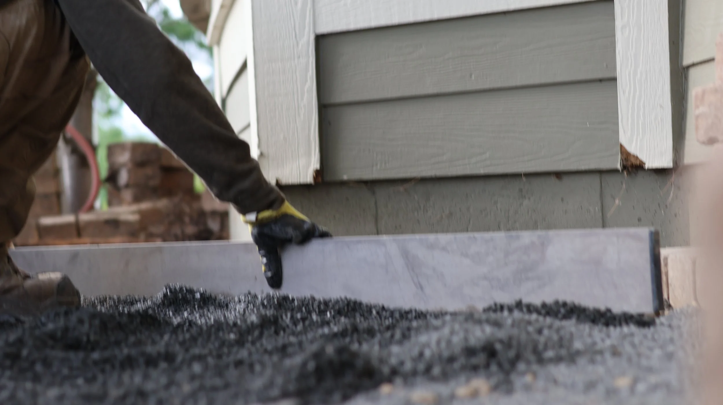 A worker wearing gloves and a brown jacket pouring hot asphalt to lay a new driveway near a house with siding.