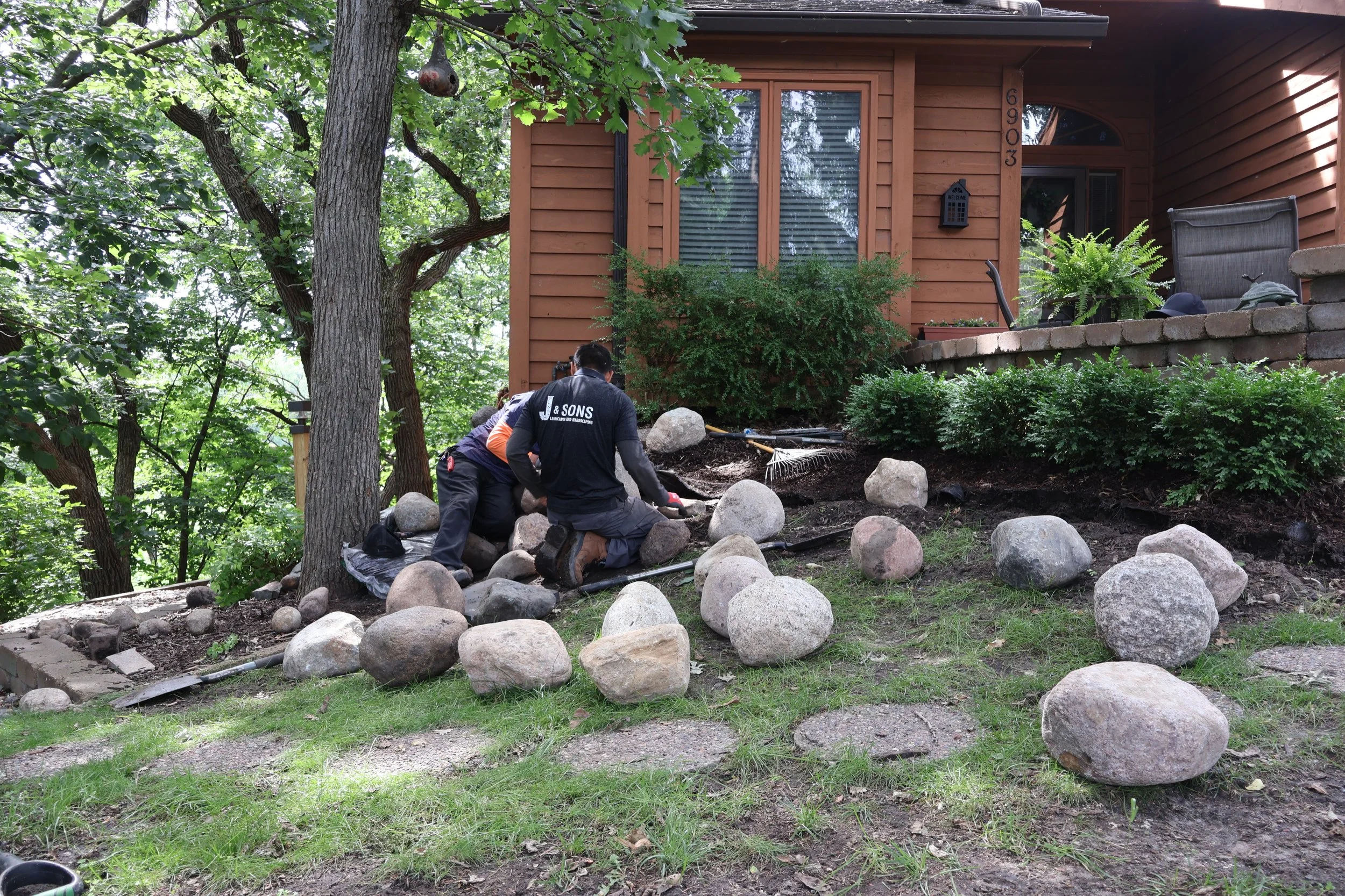 Two workers installing large rocks on a sloped yard near a brown house with green trees and shrubs.