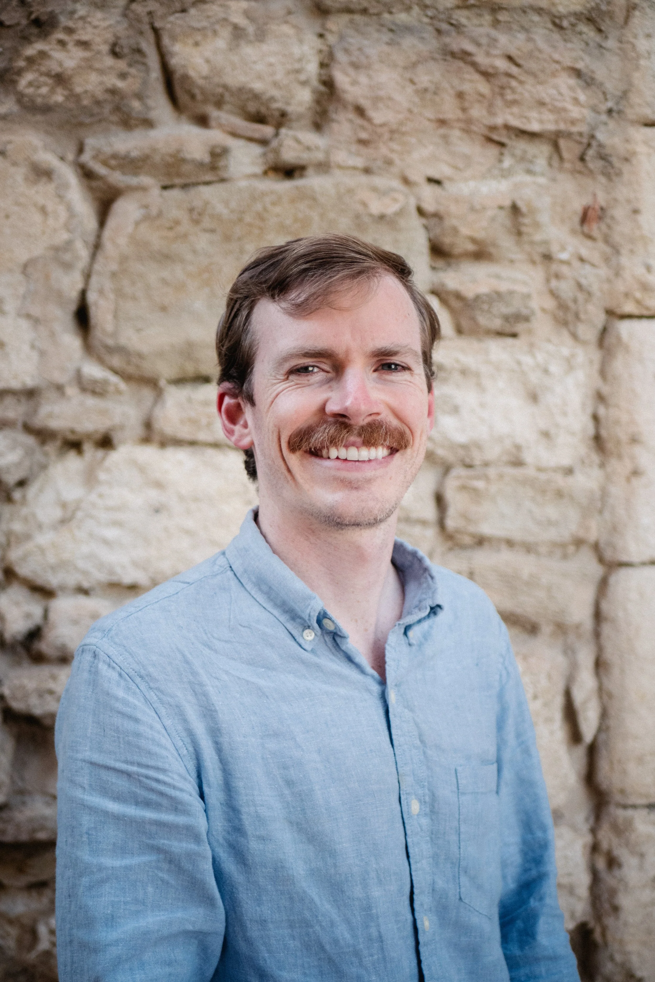A man with a light blue button-up shirt and a mustache smiling in front of a stone wall.