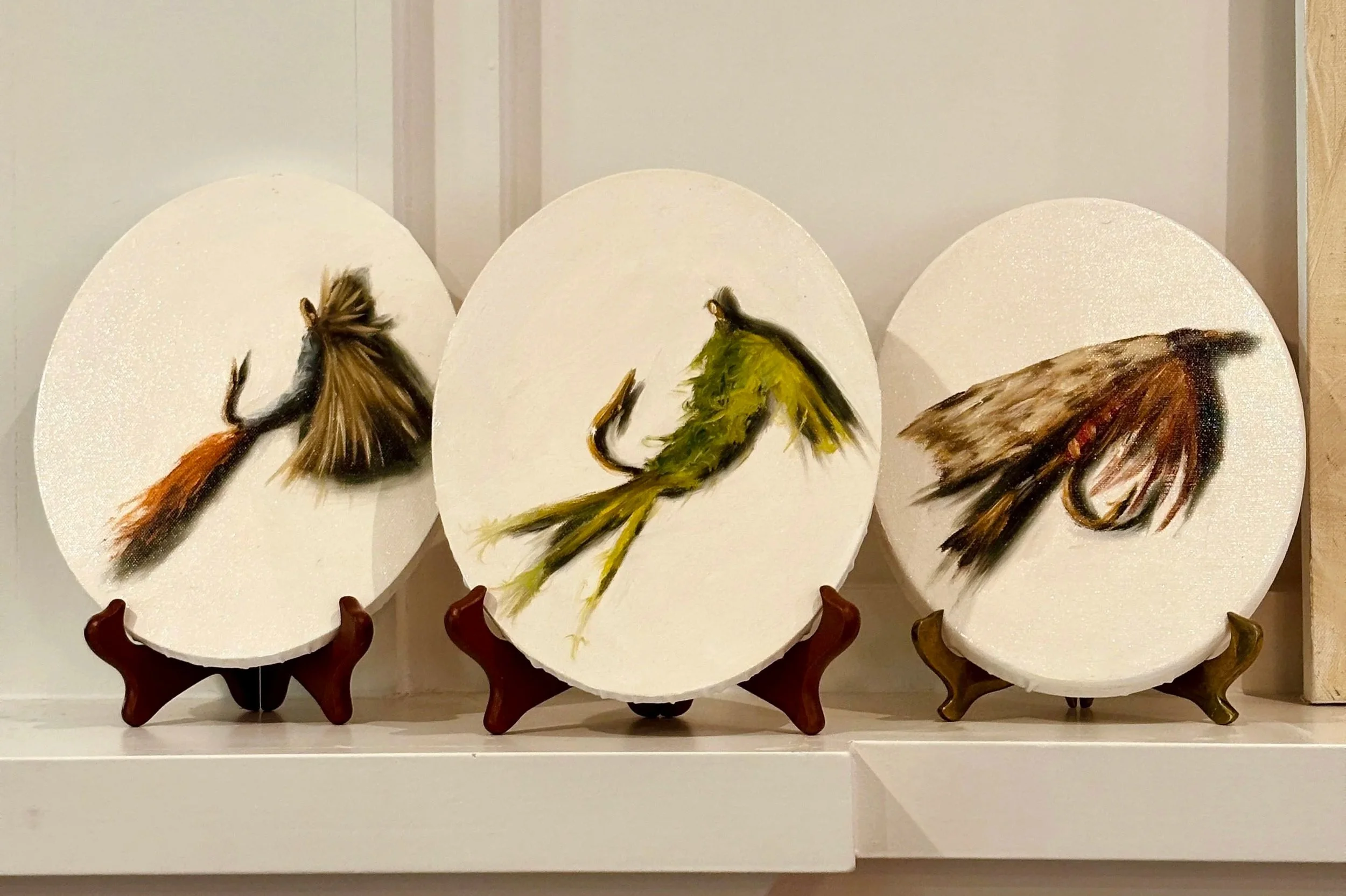 Three small oval oil paintings of fly fishing lures by Hampton Watts, displayed on wooden easels — detailed studies of classic wet flies painted in earth tones and olive greens