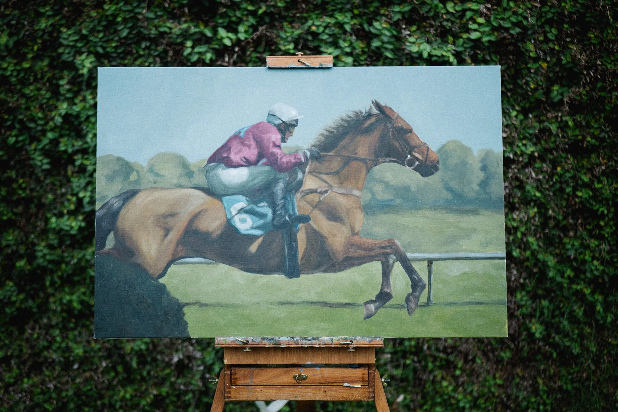 Aiken in April by Hampton Watts, equestrian oil painting on easel against green ivy, steeplechase horse and jockey clearing a jump