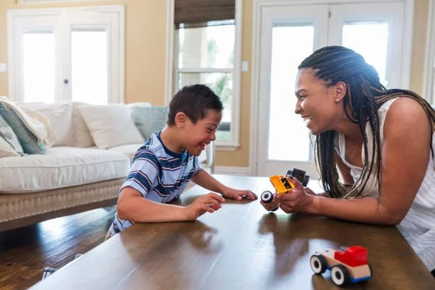 A woman and a young boy playing with toy cars on a wooden table in a bright living room.