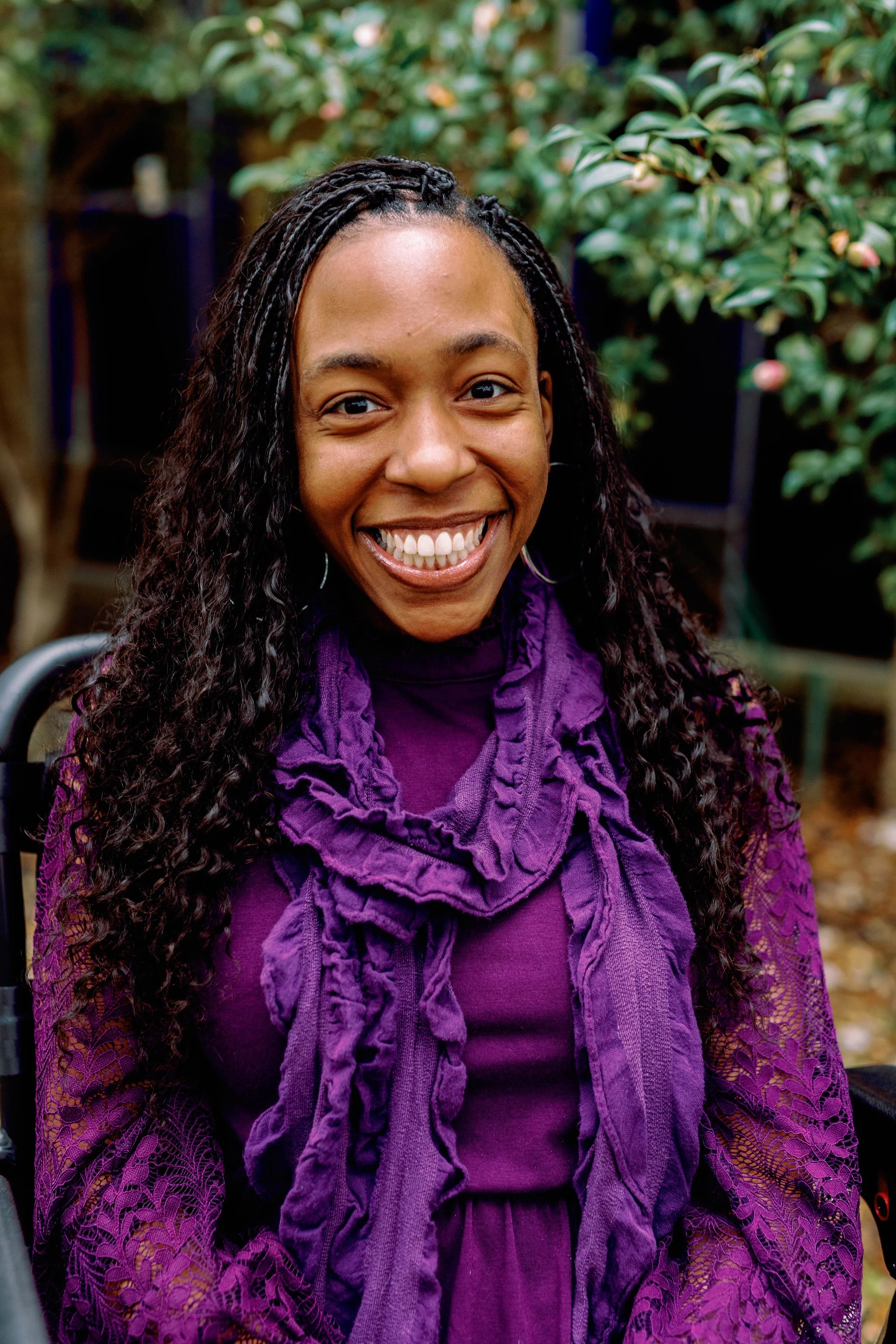 Smiling woman with long curly hair wearing a purple dress and matching scarf, sitting outdoors surrounded by greenery.