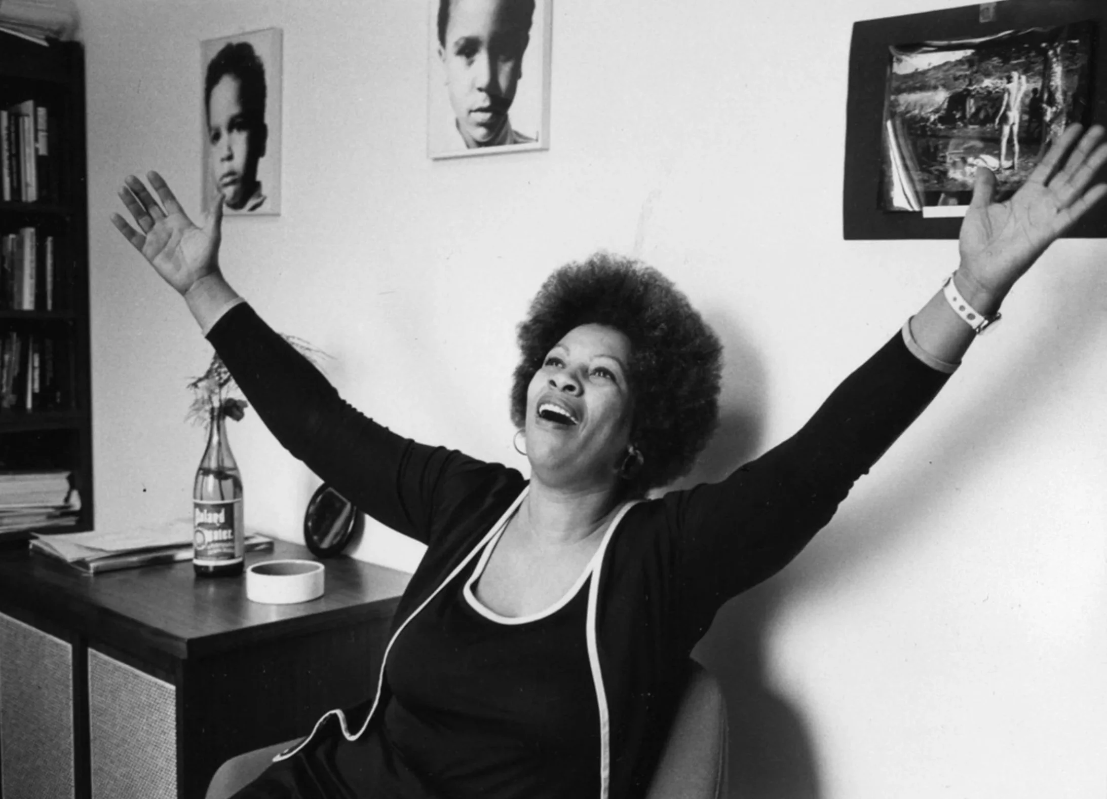 Toni Morrison joyfully raising her arms with a wide smile in a room decorated with wall art, books, and a table with a bottle and a cup.