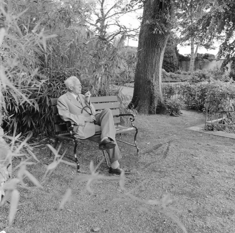 Carl Jung sitting on a park bench, smoking a pipe, surrounded by trees and bushes in a peaceful garden setting.