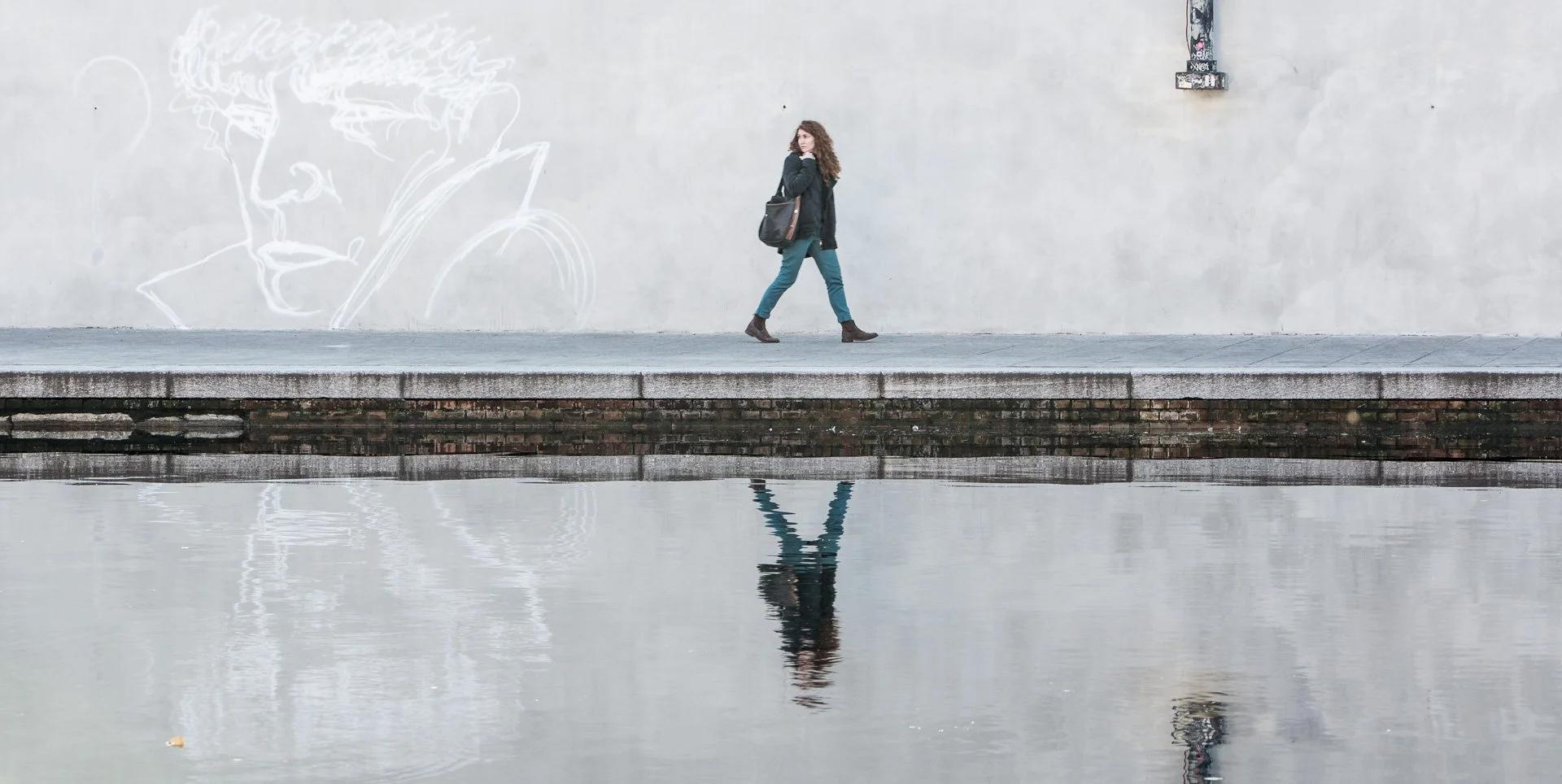 A woman with long curly hair, dark coat, and teal pants walking along a sidewalk next to a body of water, with her reflection visible in the water. She is carrying a black bag and appears to be looking at the camera. There is a mural of a face outline on the wall behind her.