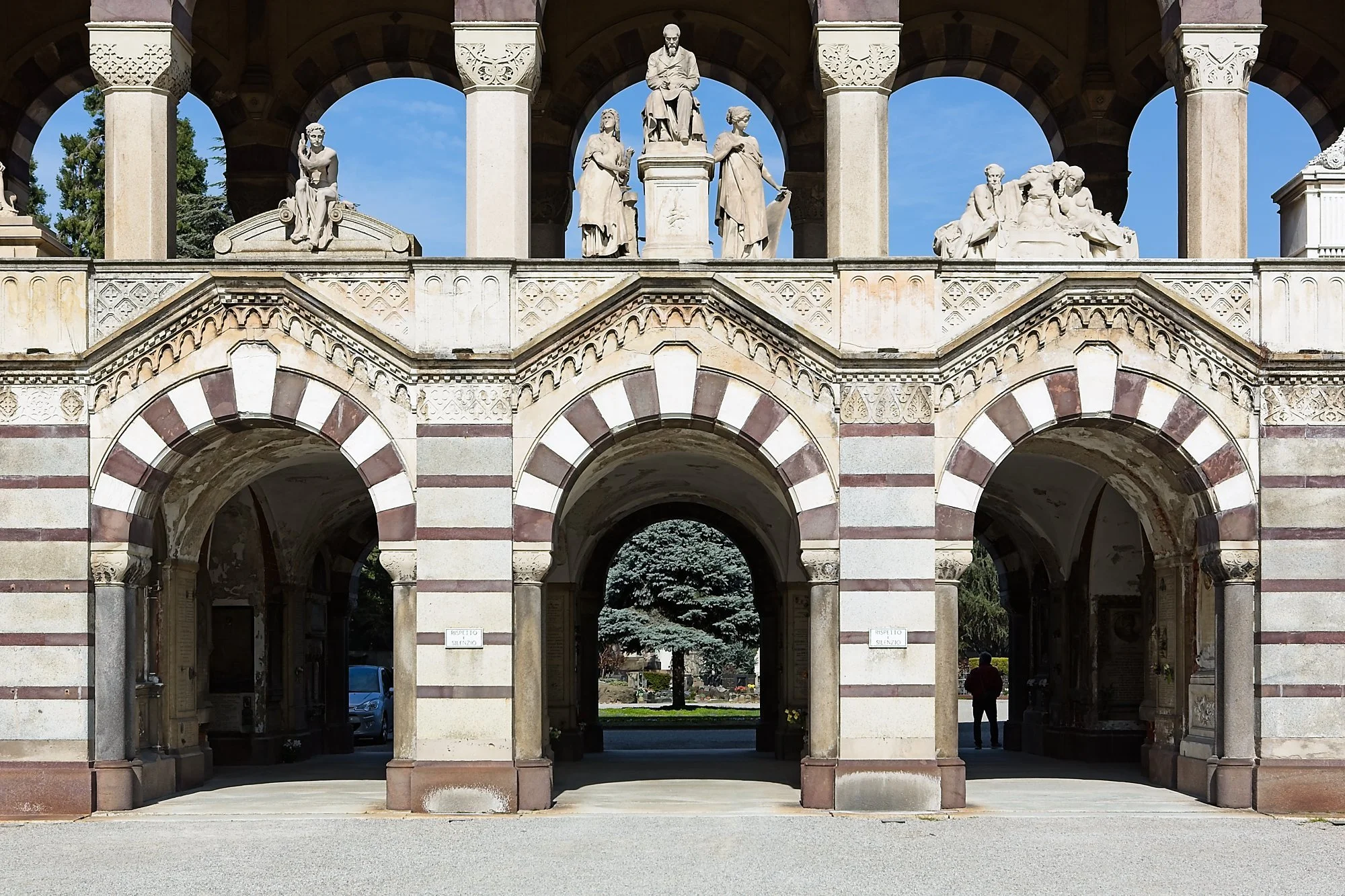 The image shows a historic stone structure with three arched entrances, decorated with statues on top, and a blue sky in the background. There are people seen in the distance inside the structure.