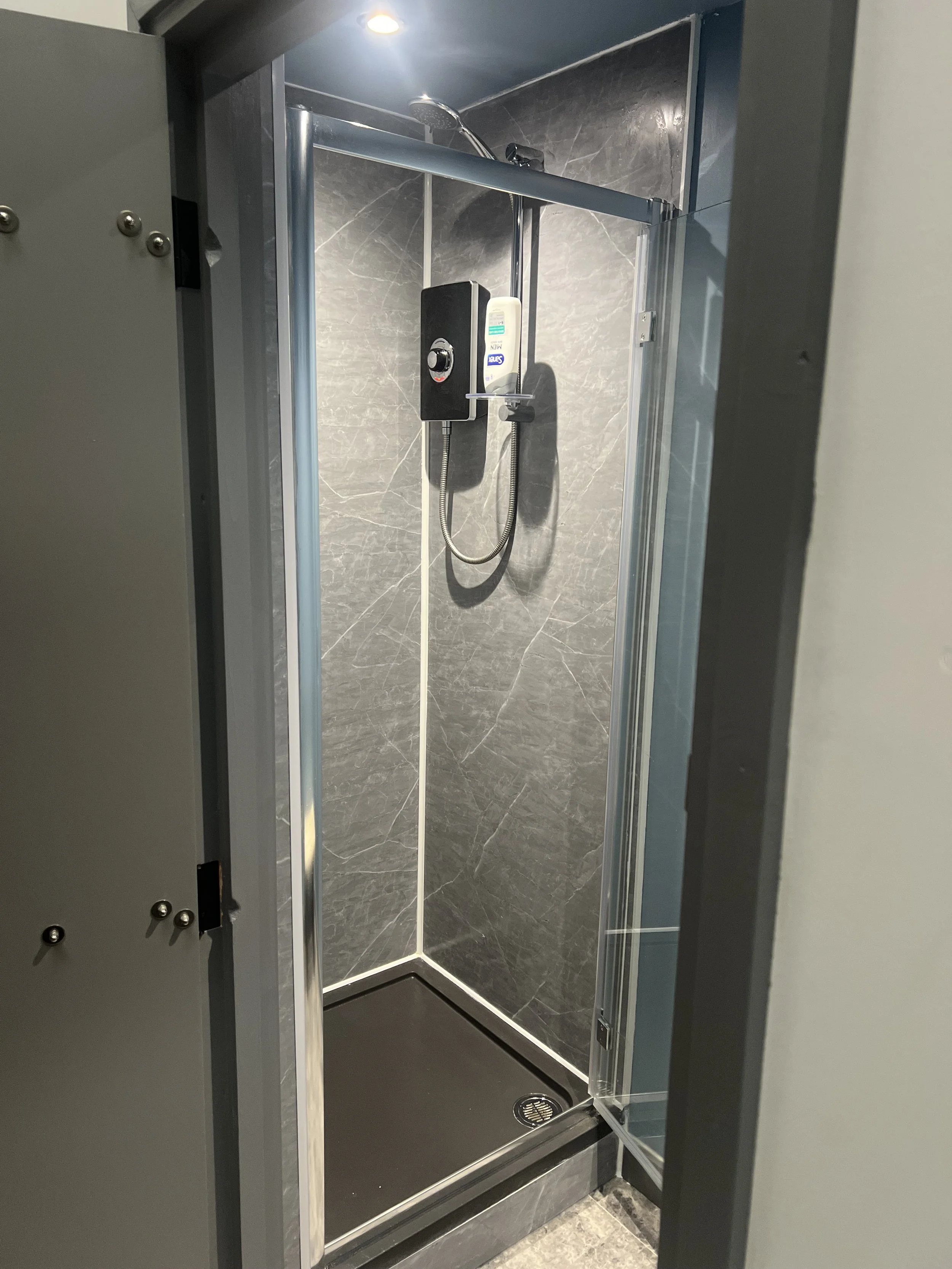 Modern glass-enclosed shower with marble-look gray tile walls, rainfall showerhead, handheld sprayer, and soap dispenser