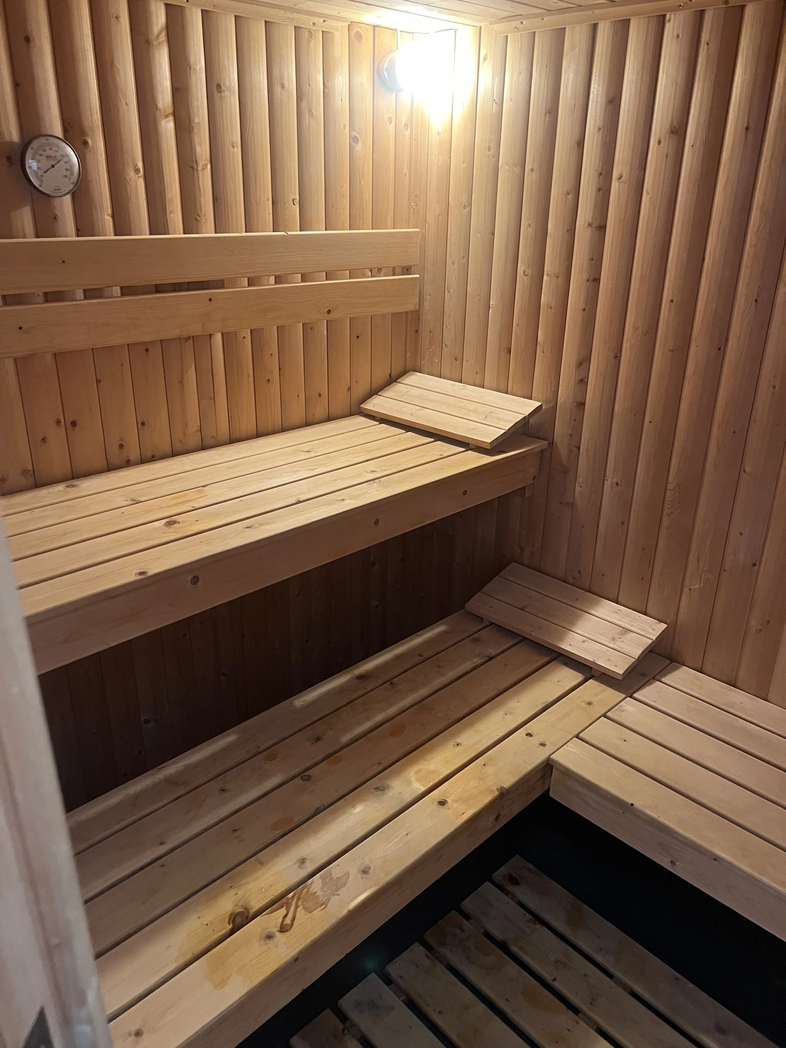 Wooden sauna interior with tiered benches, a bright wall light, and a round temperature gauge.