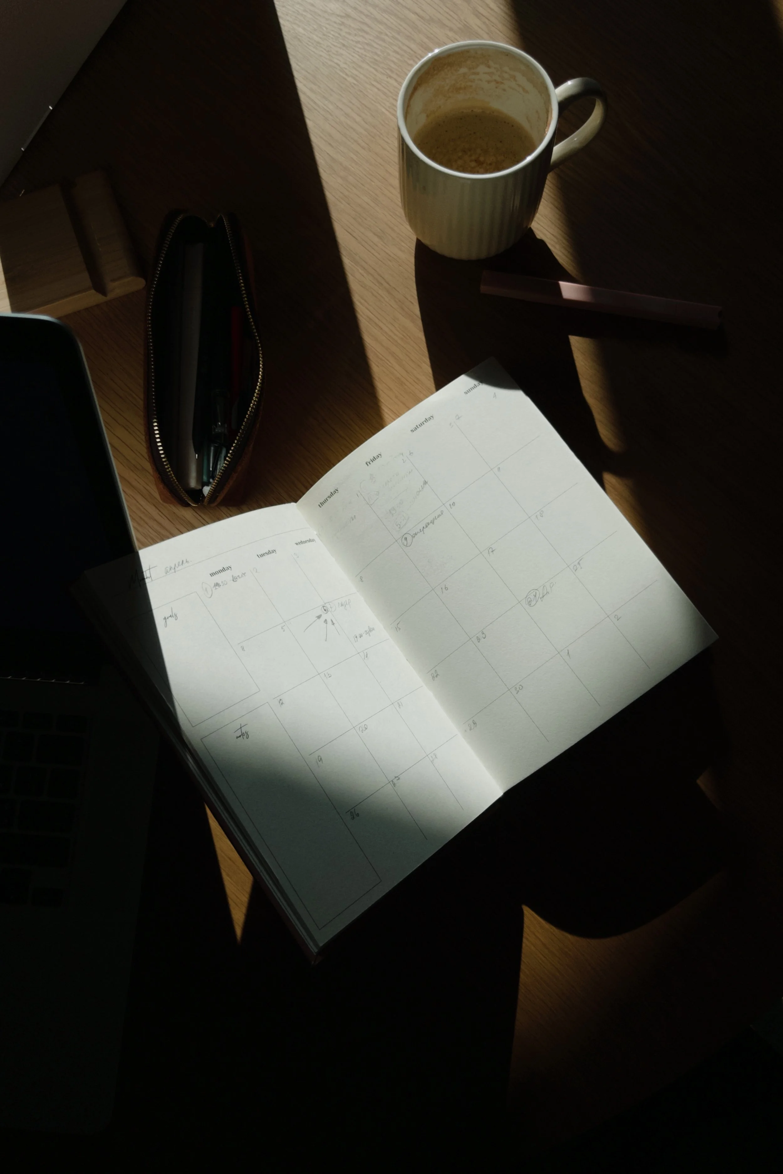 A desk with a paper planner, a pen in a zippered pouch, a coffee mug with leftover coffee, and a laptop, with sunlight casting shadows.