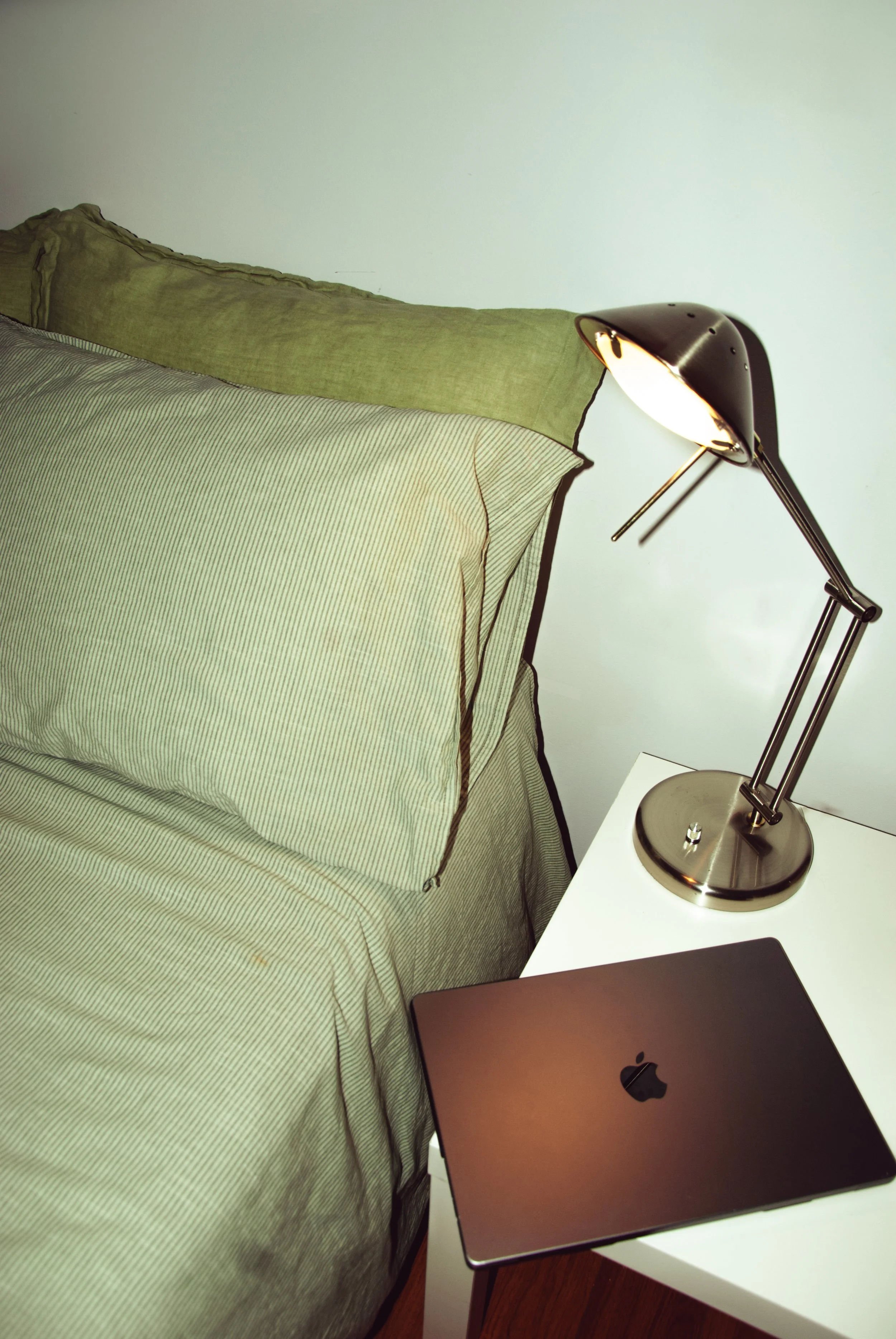 A bedroom with a bed covered in striped and solid-colored bedding, a green pillow, a black desk lamp, an Apple MacBook on a white bedside table, and a smartphone on the table against a white wall.
