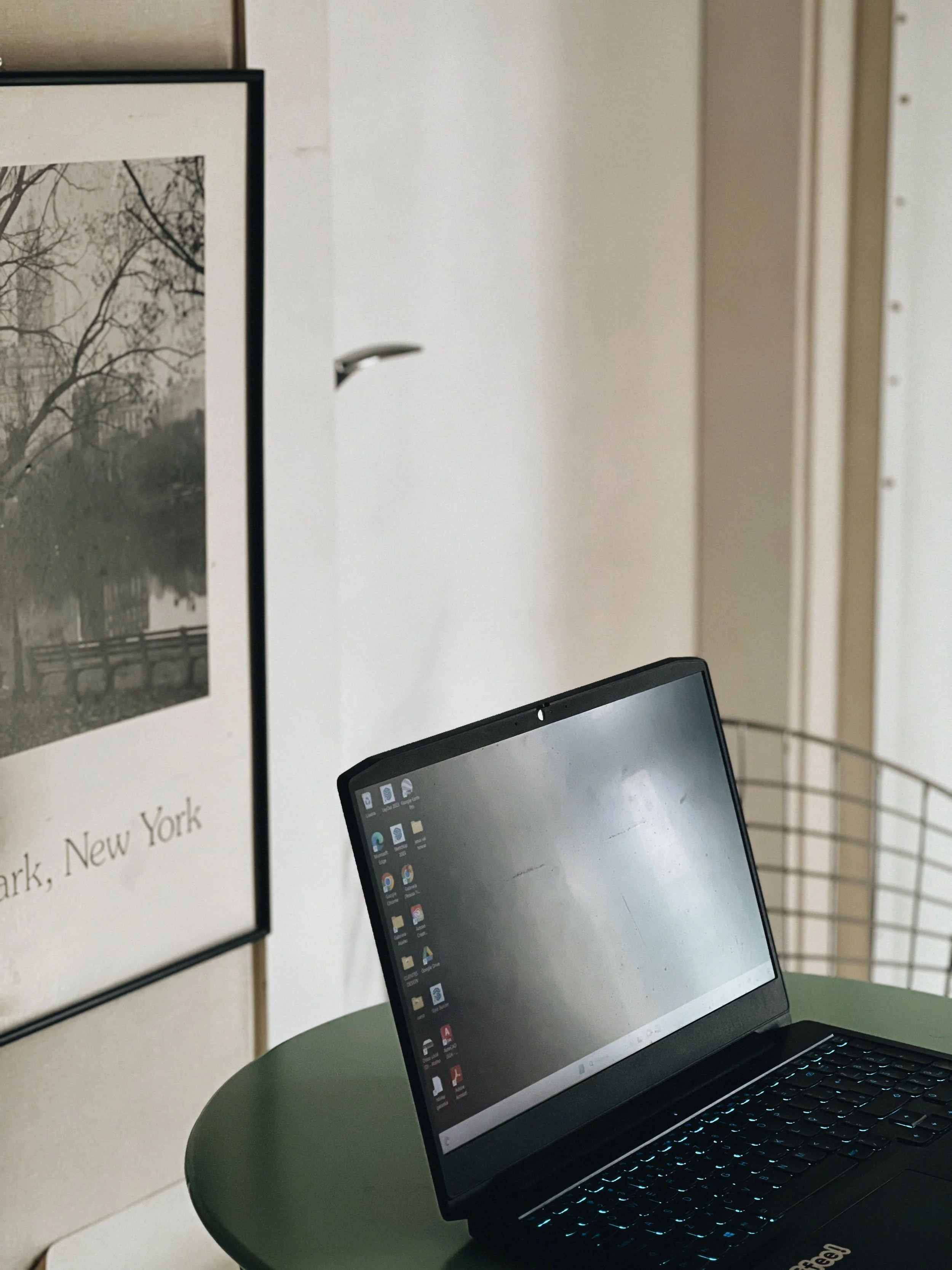 A black laptop with a glowing keyboard on a green round table, with a white wall and a framed black-and-white photograph of a tree-lined river or canal in the background.