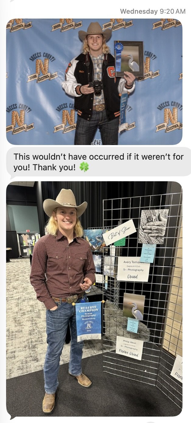 A person dressed in a cowboy hat, plaid shirt, and jeans holding a camera and a framed bird photo standing in front of a blue backdrop with 'Nueces County Junior Livestock Show' logos. Another person in a cowboy hat, plaid shirt, jeans, and boots standing beside a display board with award signs, and a photo of a bird.