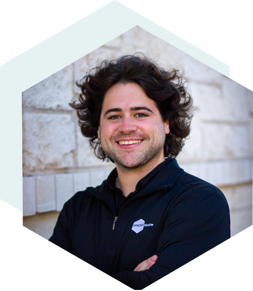 Smiling person with curly dark hair wearing a black zip-up jacket, standing with arms crossed in front of a light stone wall, framed in a hexagonal shape.