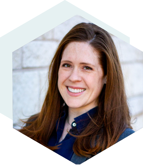 Smiling person with long brown hair wearing a blue shirt, standing in front of a light stone wall, framed in a hexagonal shape.
