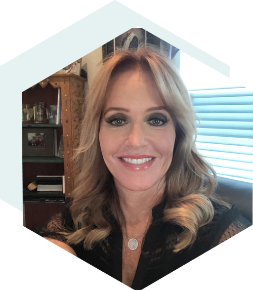 Smiling person with blonde shoulder-length hair wearing a black top, indoors with shelves and a window in the background, framed in a hexagonal shape.