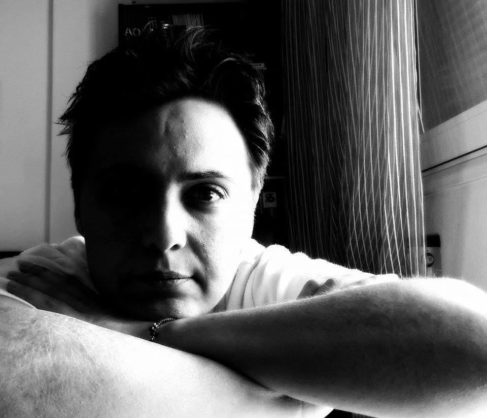 Black and white photo of a person resting their head on crossed arms by a window with vertical blinds, softly lit from the side.