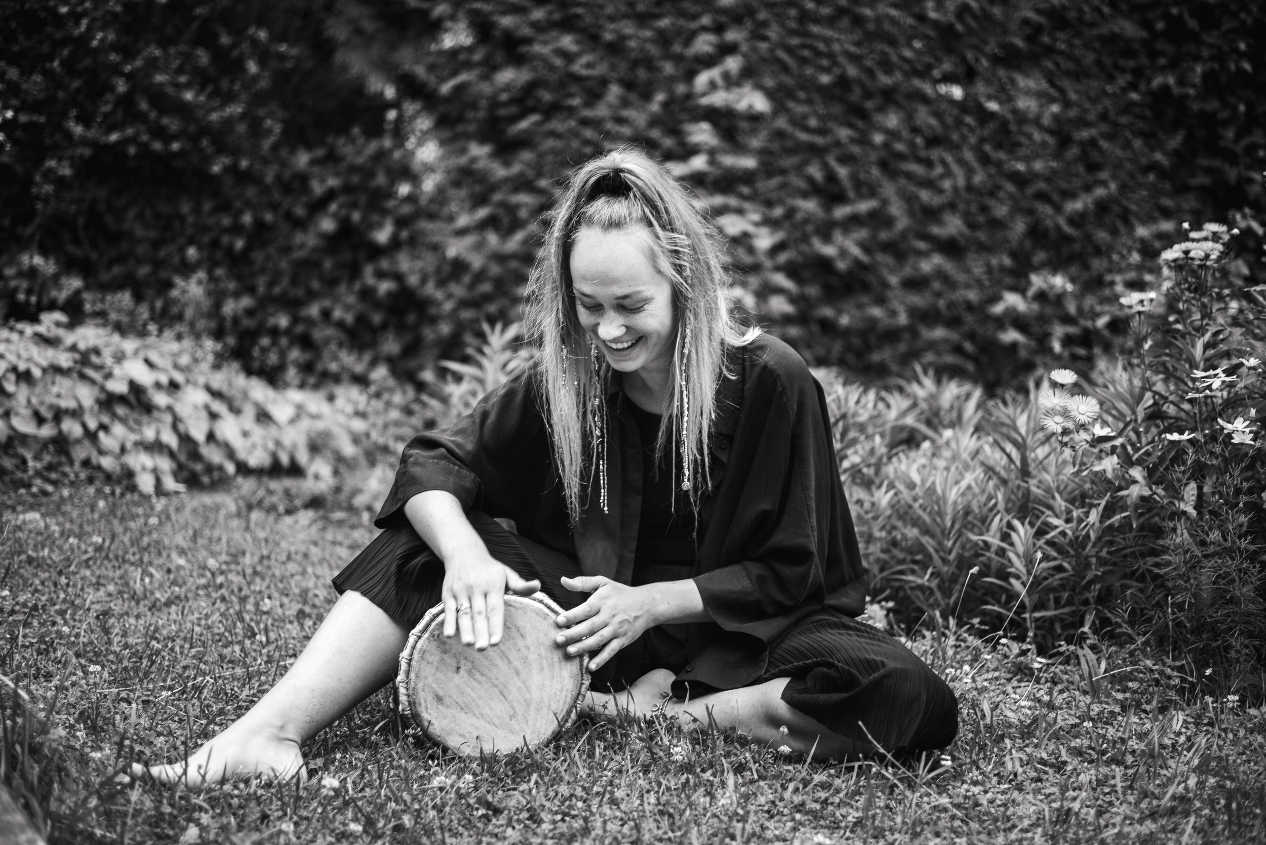 Katharina Wirth sitting in nature, smiling and playing the drum / Djembé. Black & White Colors. Sexological Bodytherapy.