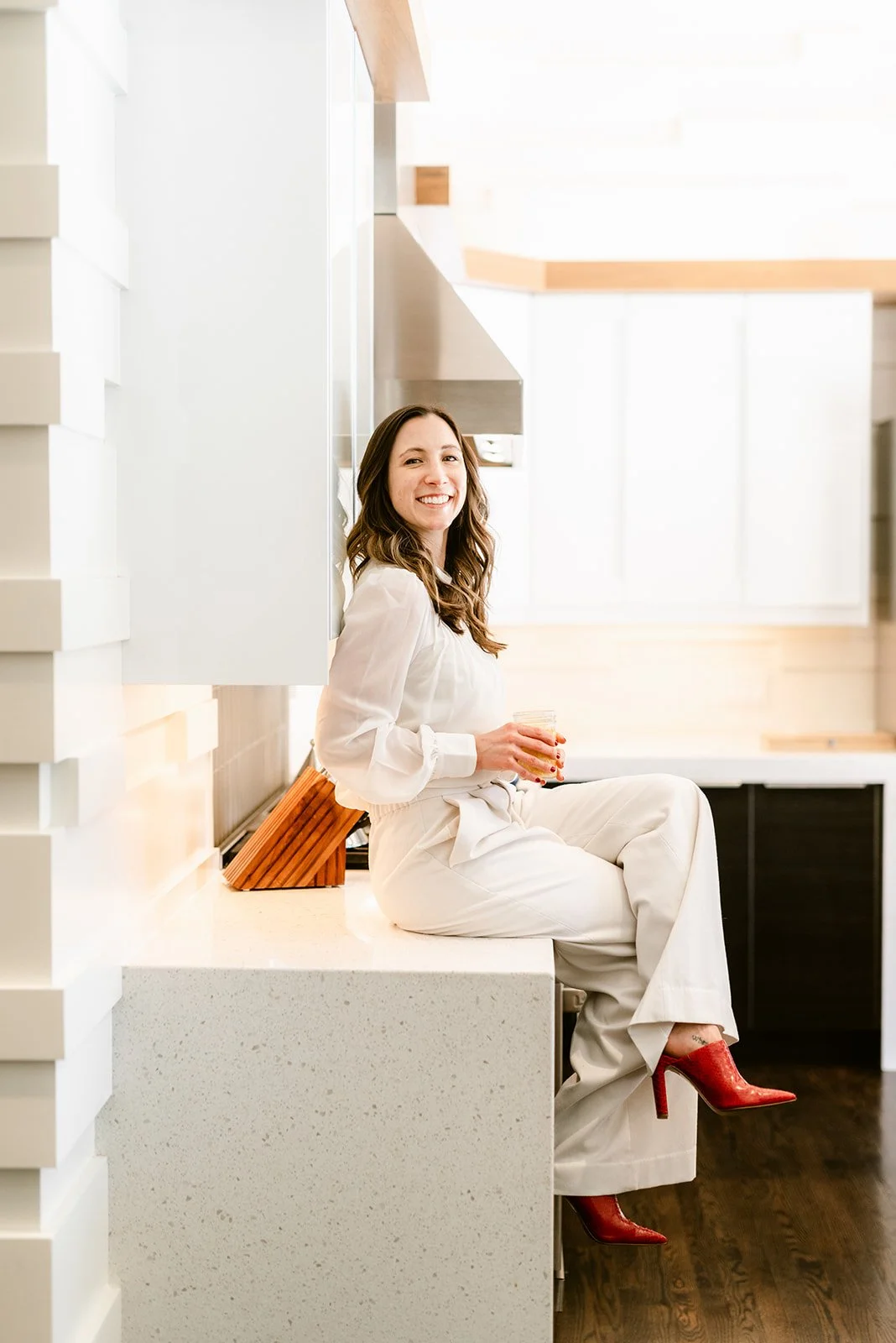Woman in white outfit sitting on kitchen counter, holding a drink, wearing red high heels, modern kitchen.