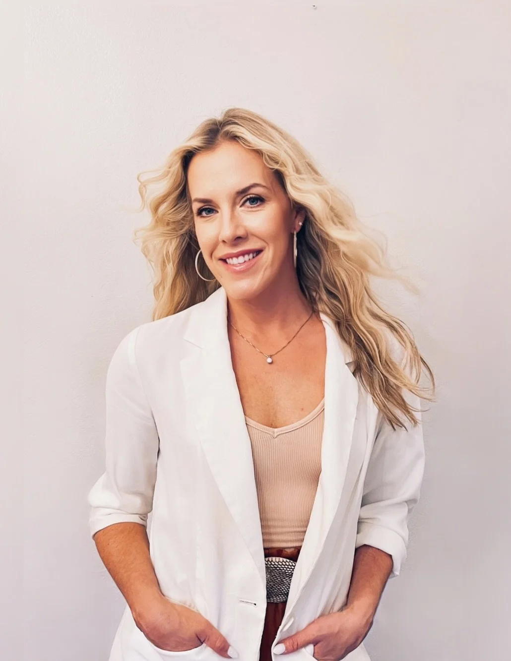 A woman with blonde curly hair smiling, wearing a white blazer, beige top, hoop earrings, and a necklace, standing against a plain white wall.