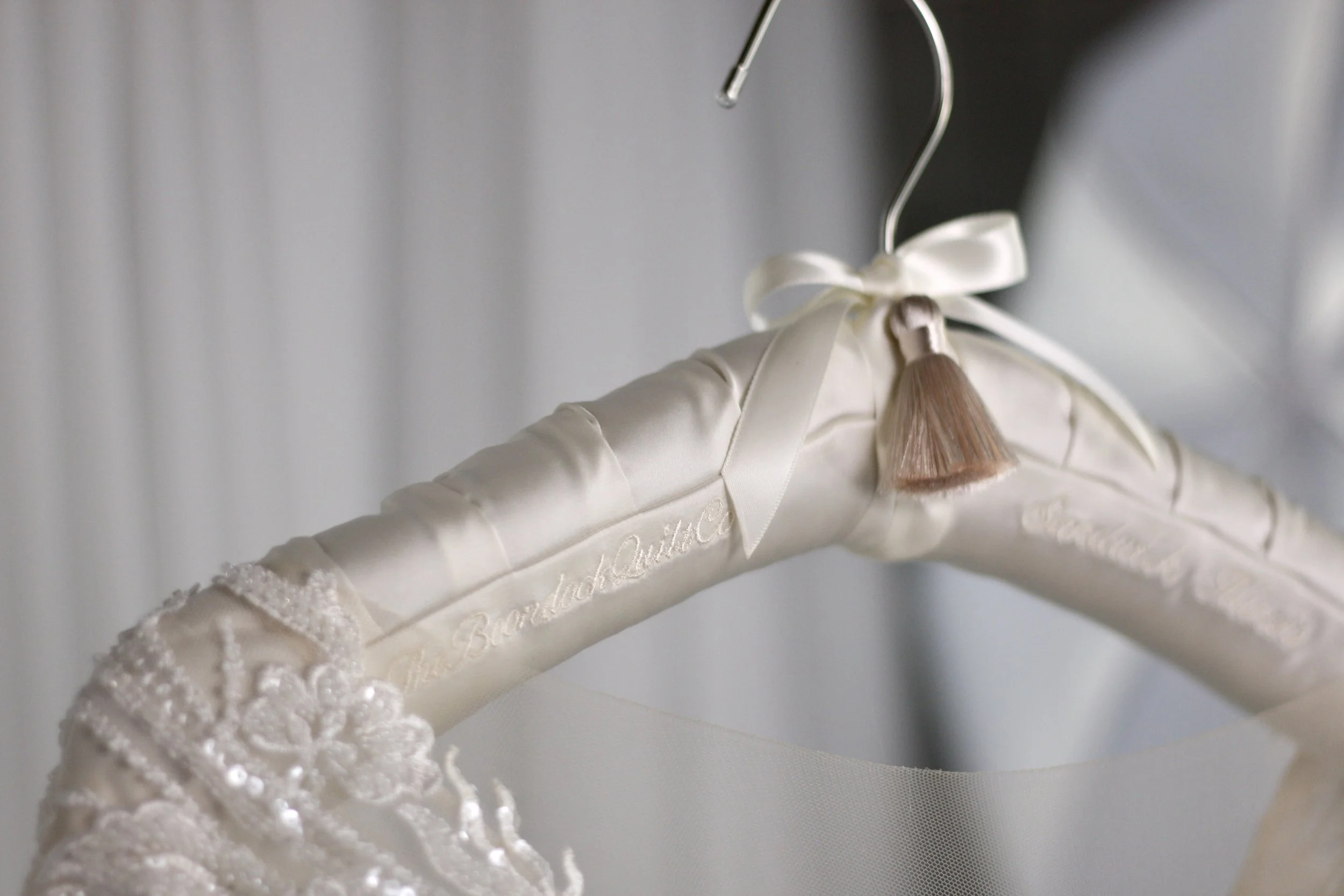 Close-up of an elegant, embroidered wedding dress on a hanger with a satin ribbon and a small brown tassel.