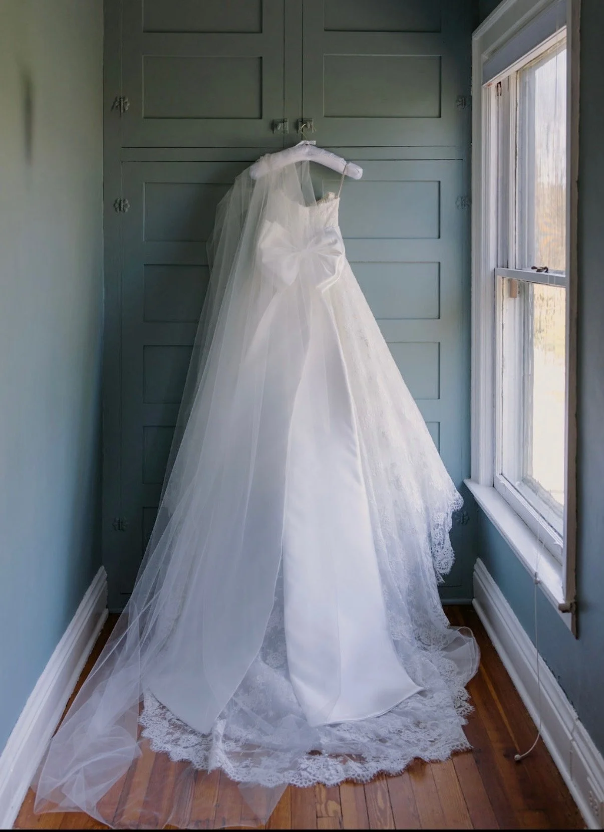 A white wedding dress with a veil hanging on a personalized satin bridal hanger, placed against a wall near a window in a room with light blue walls and wood flooring.