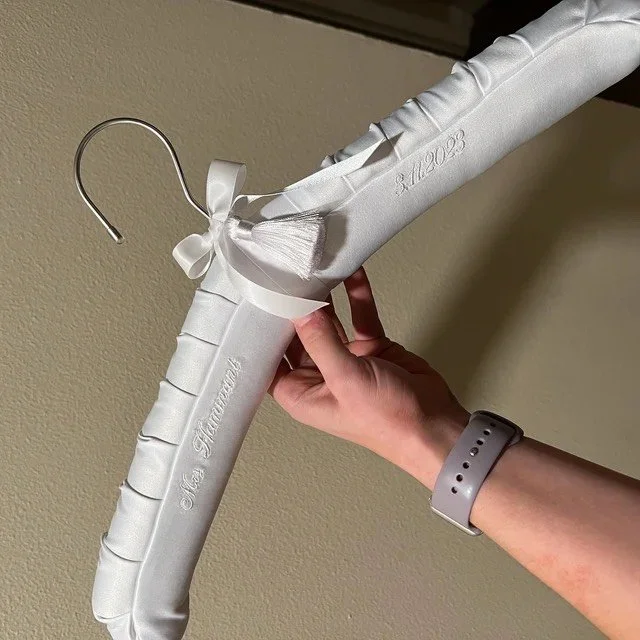 A white satin hanger shaped like a large bouquet or decorative arrangement, with a satin ribbon and bow, being held in a person's hand against a plain wall.