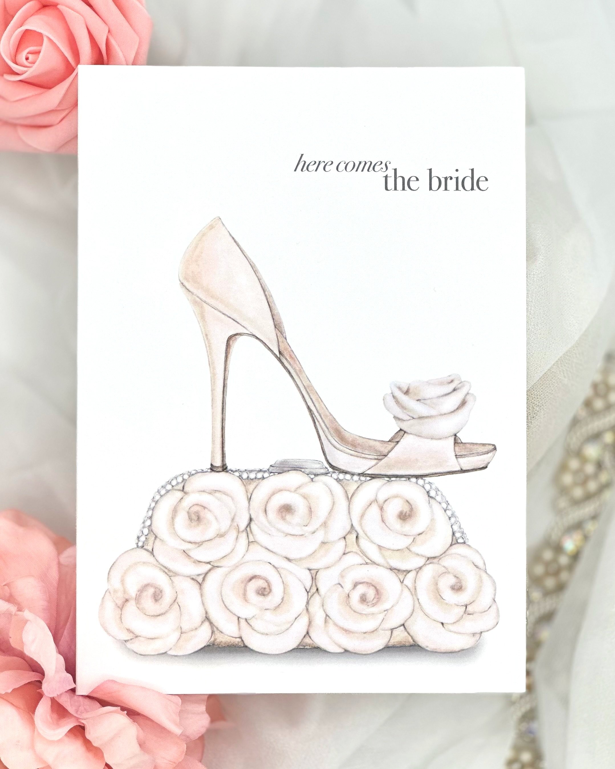 Wedding card featuring glamorous high-heeled shoes adorned with a delicate rose, paired with a clutch purse with soft ivory pattern.