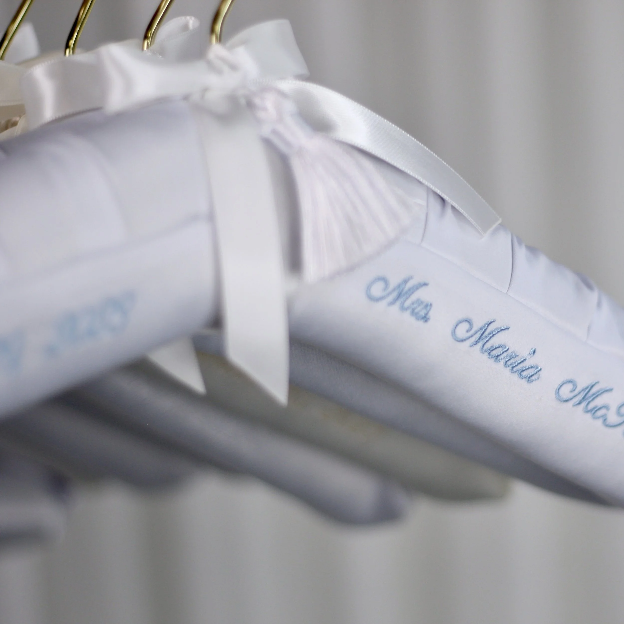 White fabric garment with blue embroidered text reading 'Mia Maial Ma...', hanging on a gold hanger with white satin ribbon and bow details.