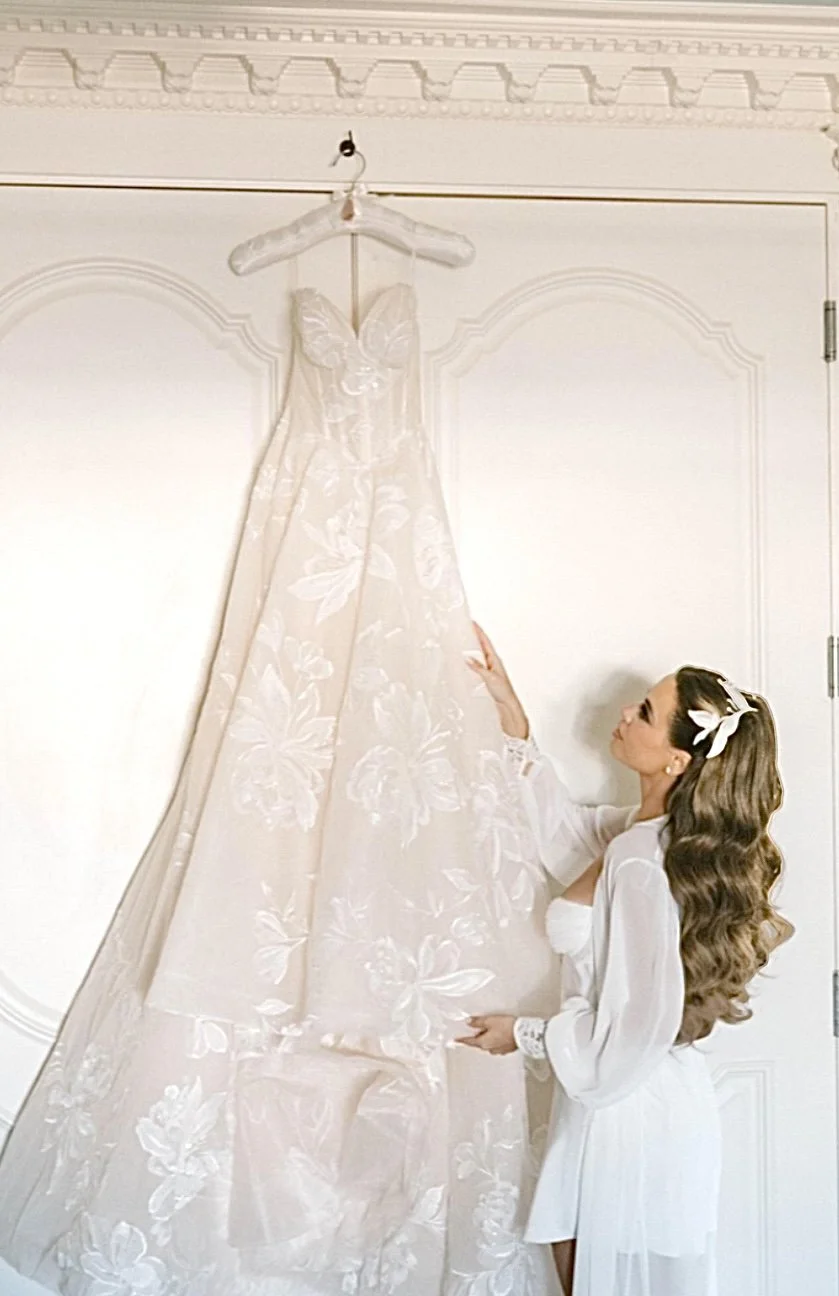 A woman in a white robe looks at a long white wedding dress with floral embroidery, hanging on a hanger on a white wall.