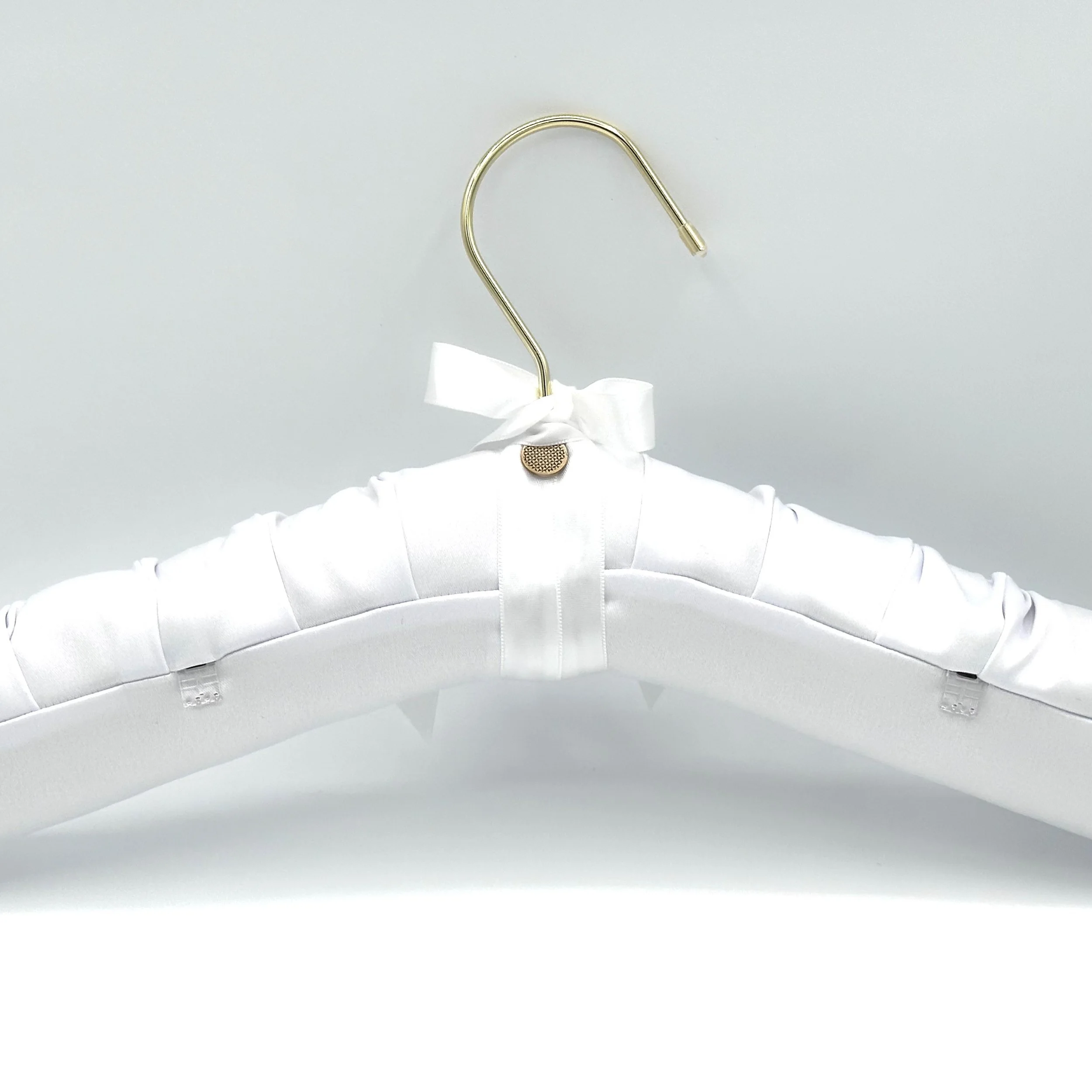 A white satin wedding dress hanger with a bow, hanging on a gold hook against a gray background.