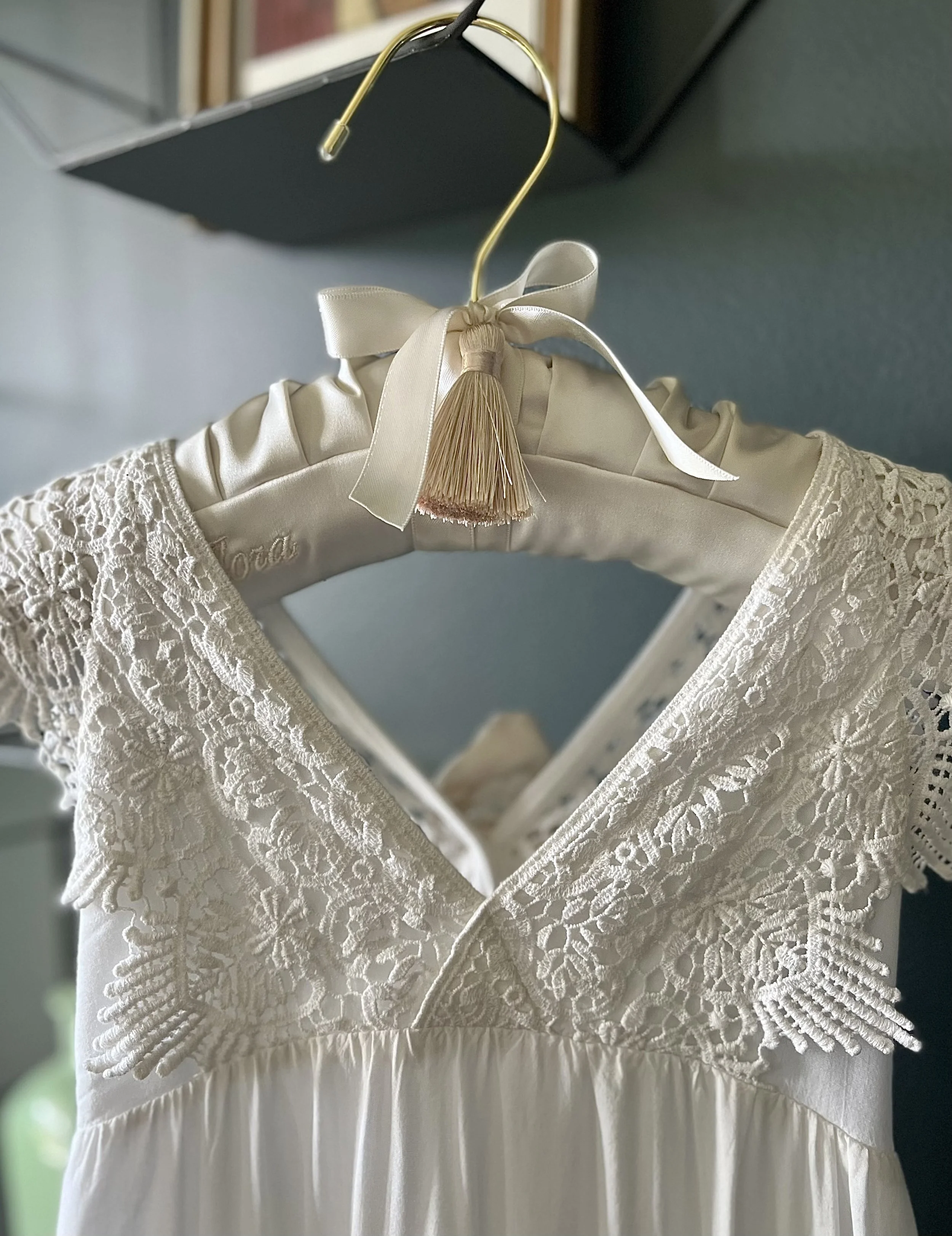 A white lace dress hanging on a satin padded hanger with a ribbon and tassel embellishment.