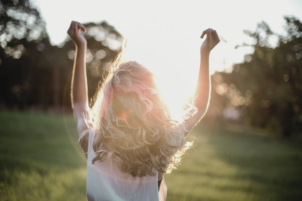 Woman with curly blonde hair raising her arms in a field during sunset