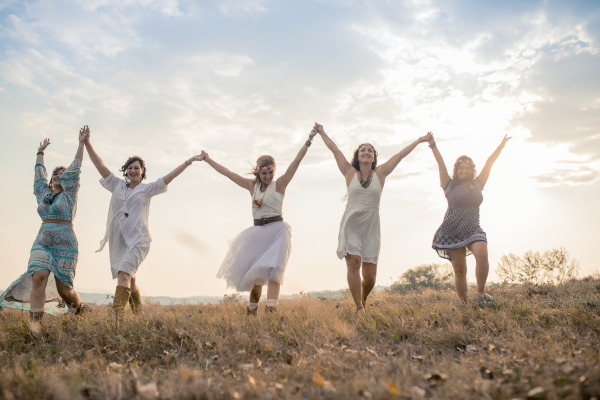 Five women holding hands and dancing outdoors in a grassy field during sunset.