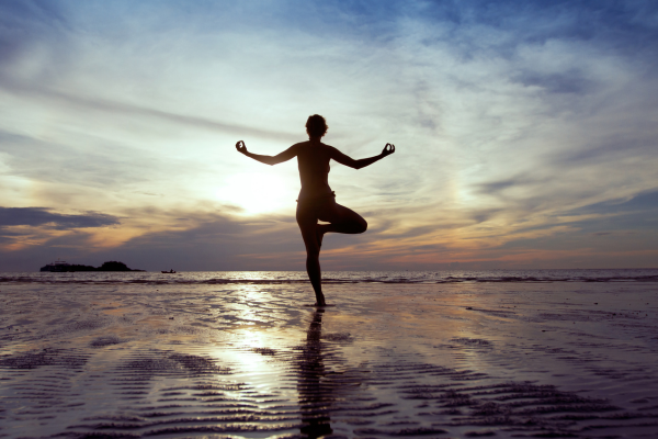 Person practicing yoga or meditation on a beach at sunset, balancing on one leg with arms raised and hands in mudras.