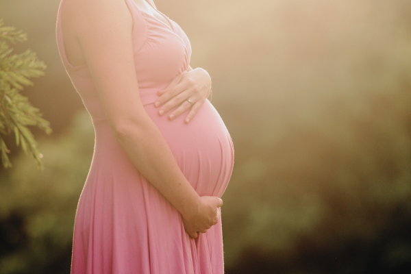 Close-up of a pregnant woman wearing a pink dress, gently holding her belly outdoors in soft sunlight.