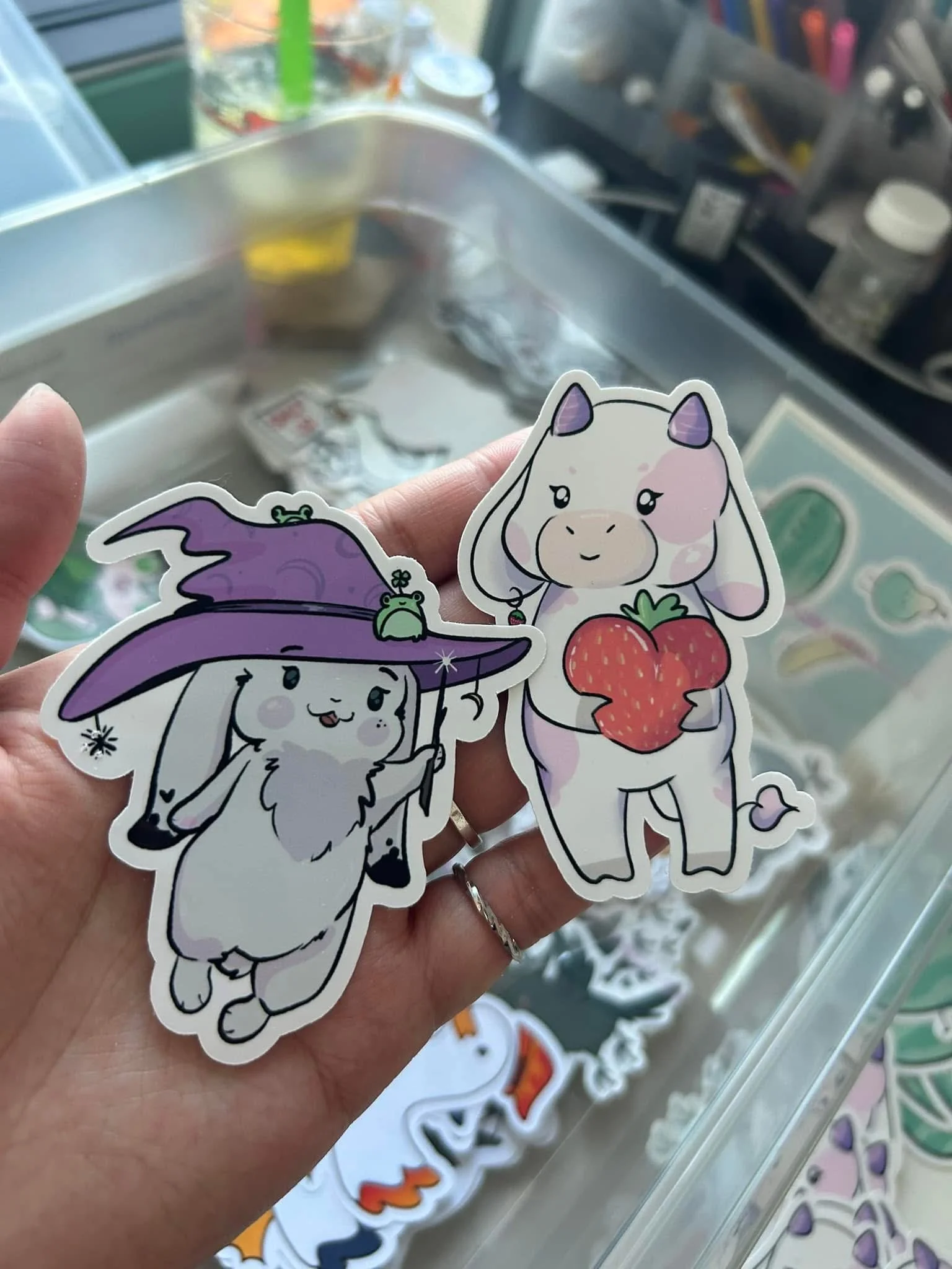 Two cartoon-style stickers: one of a cute white goat dressed as a wizard with a purple hat and tiny green frog, the other of a white calf holding a large strawberry. The background shows a cluttered drawer with craft supplies.