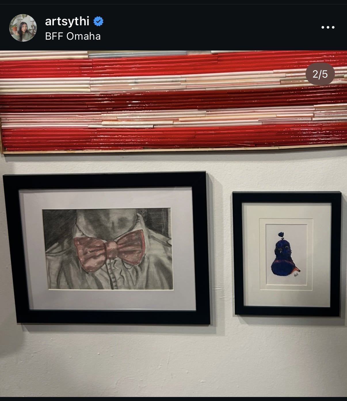 Art gallery wall displaying two framed artworks: on the left, a black and white drawing of a person wearing a red bow tie; on the right, a colorful, stylized portrait of a woman with a flower on her head.