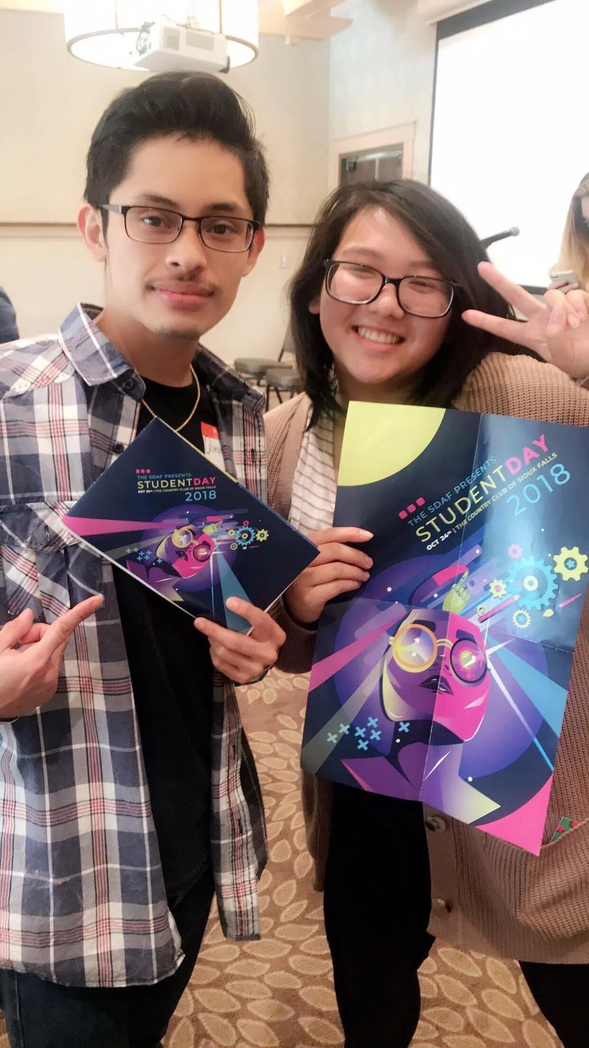 Two people holding colorful event brochures at a student event in 2018, with one making a peace sign.