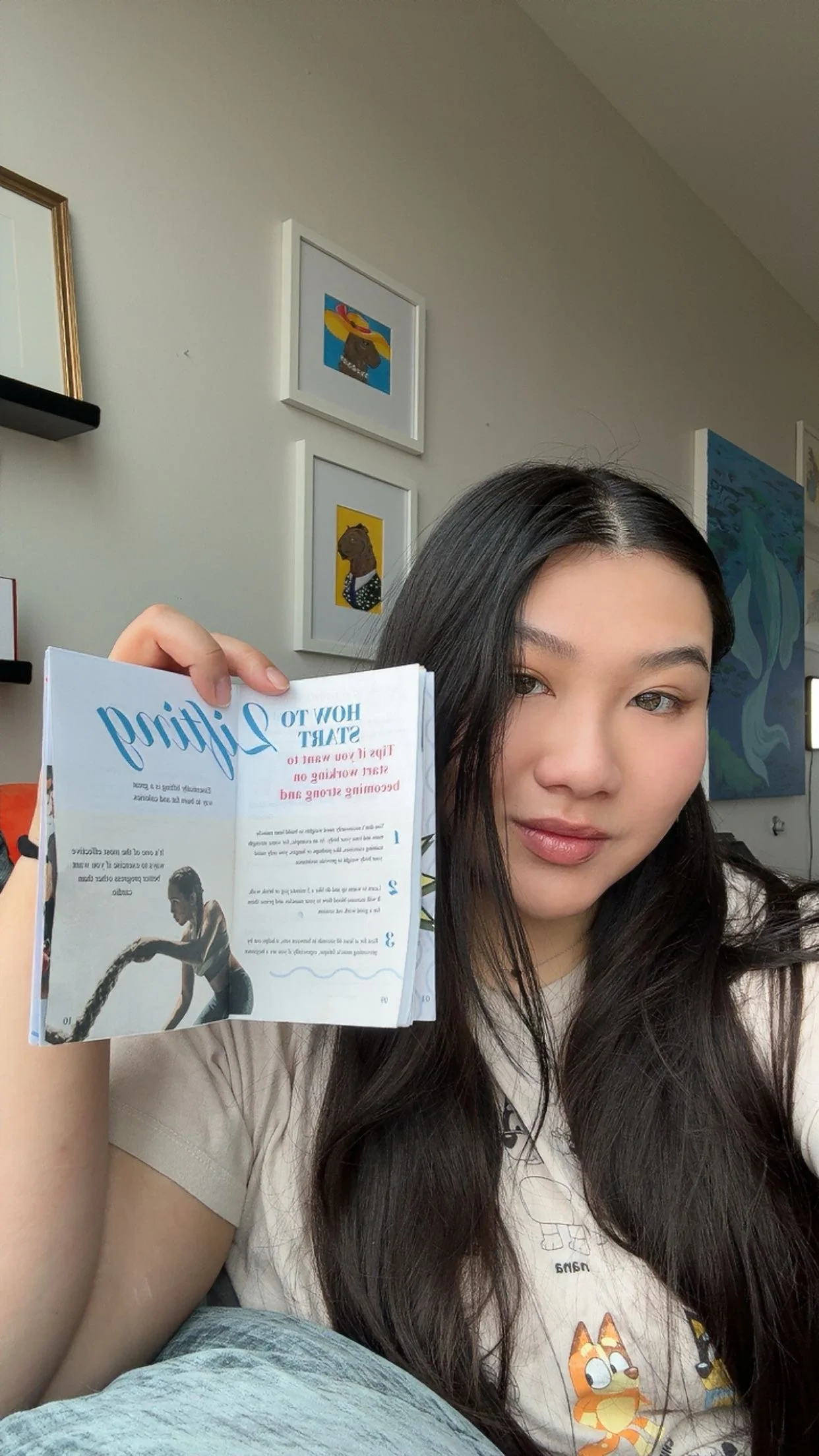 A young woman with long dark hair holding a small pamphlet or flyer, sitting in a room with framed artwork on the wall behind her.