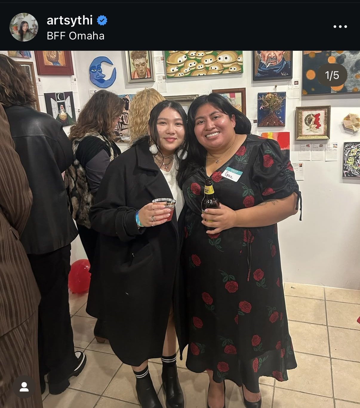 Two women smiling and holding drinks at an art gallery event, surrounded by artwork on the wall.
