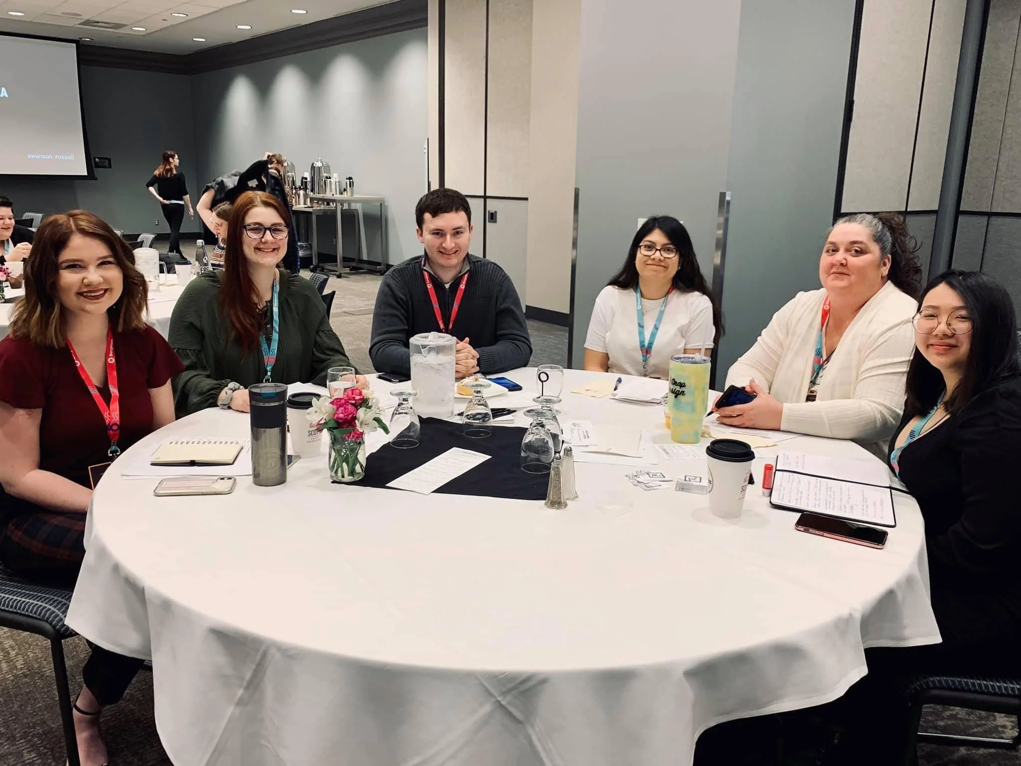 A group of seven diverse women sitting around a round table during a conference or meeting, with notebooks, pens, and drinks on the table.