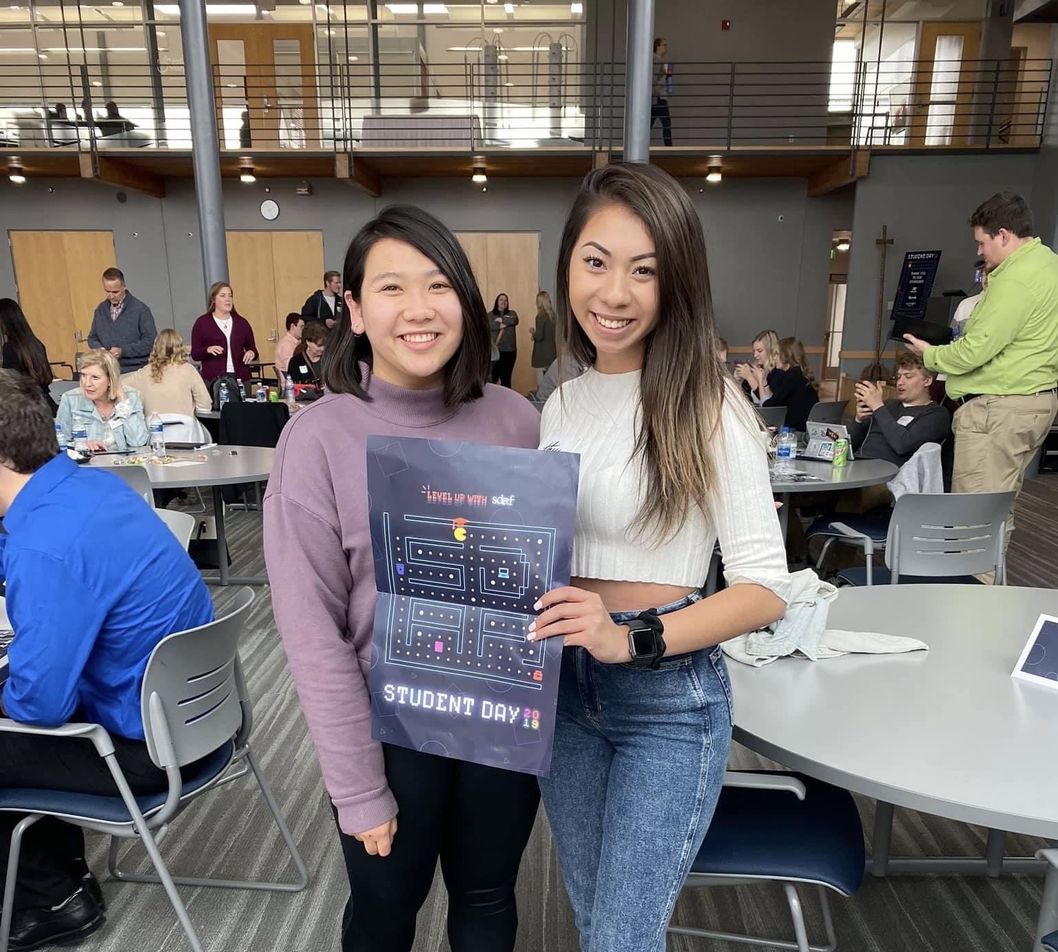 Two smiling women standing together at a gathering, one holding a poster that says 'Level Up with SLEF' and 'Student Day 2019', in an indoor event space with people sitting and standing at tables in the background.