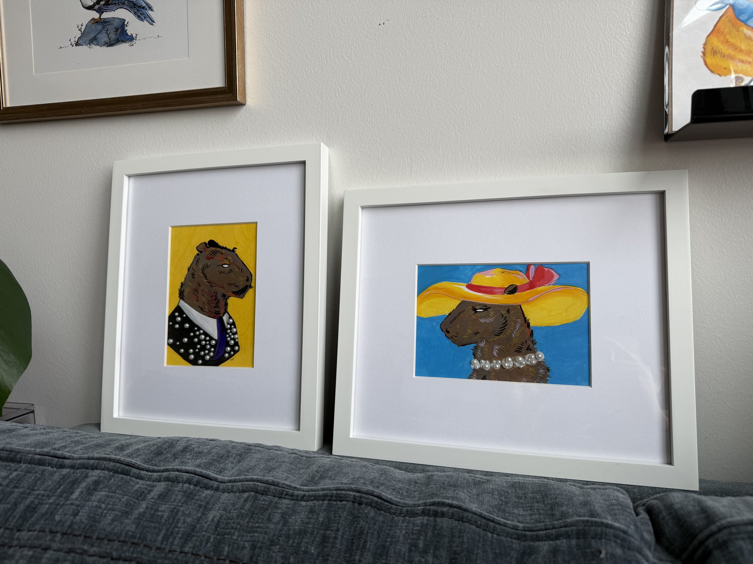 Two framed illustrations of a guinea pig dressed in fashionable accessories, one with a polka dot blazer on a yellow background, the other wearing a large sun hat and pearl necklace on a blue background, leaning against a wall.