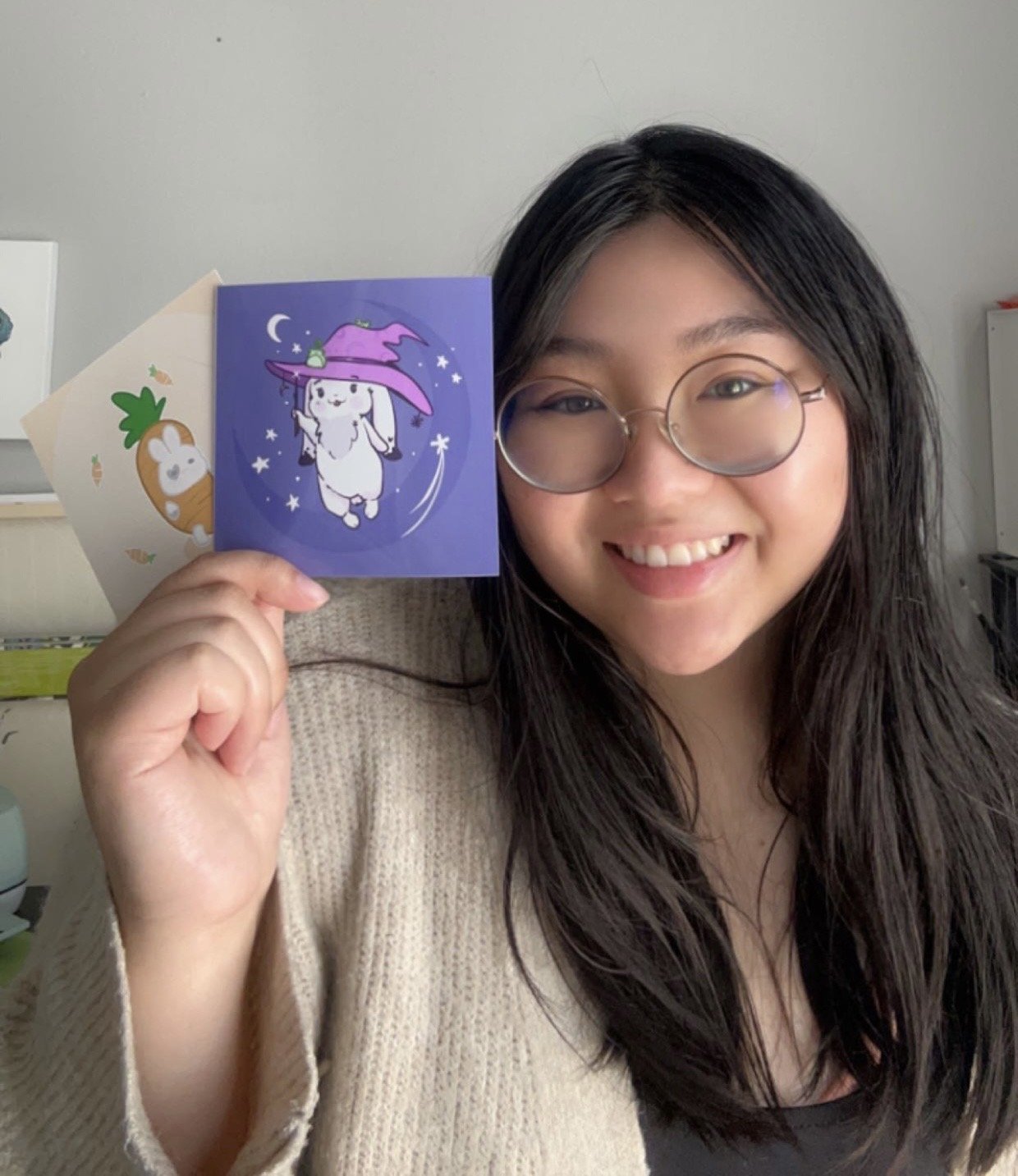 A young woman with black hair, glasses, and a beige sweater smiling and holding up two colorful stickers, one with a cute bunny and the other with a cute character wearing a purple hat.