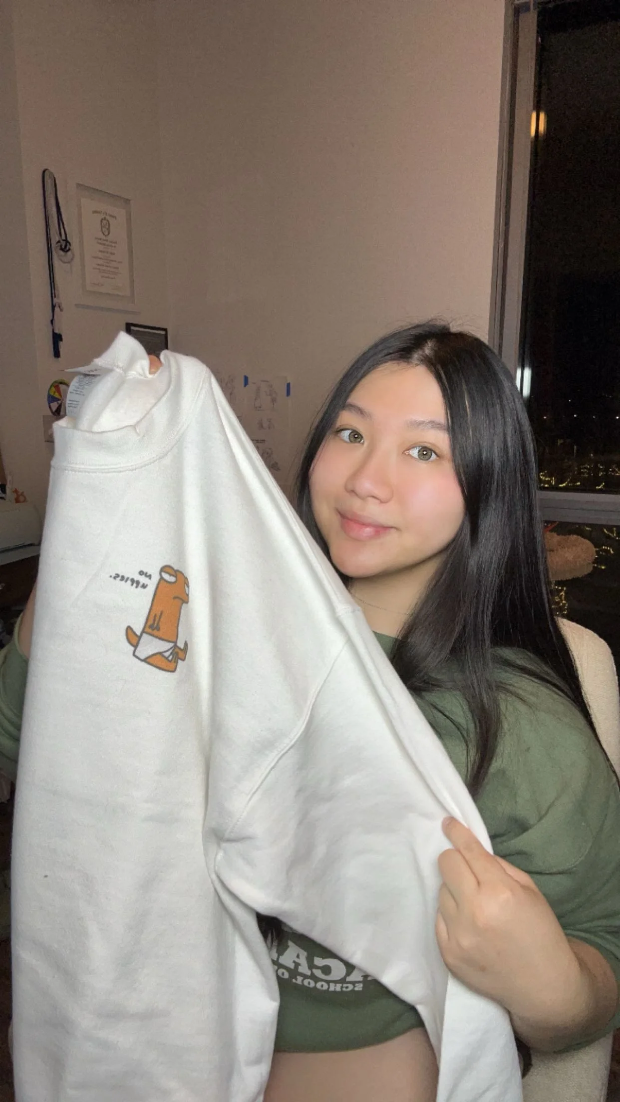 A young woman with long black hair holding a white sweatshirt with a small bear illustration and the word "WIP" on it, standing in a room with a window at night.