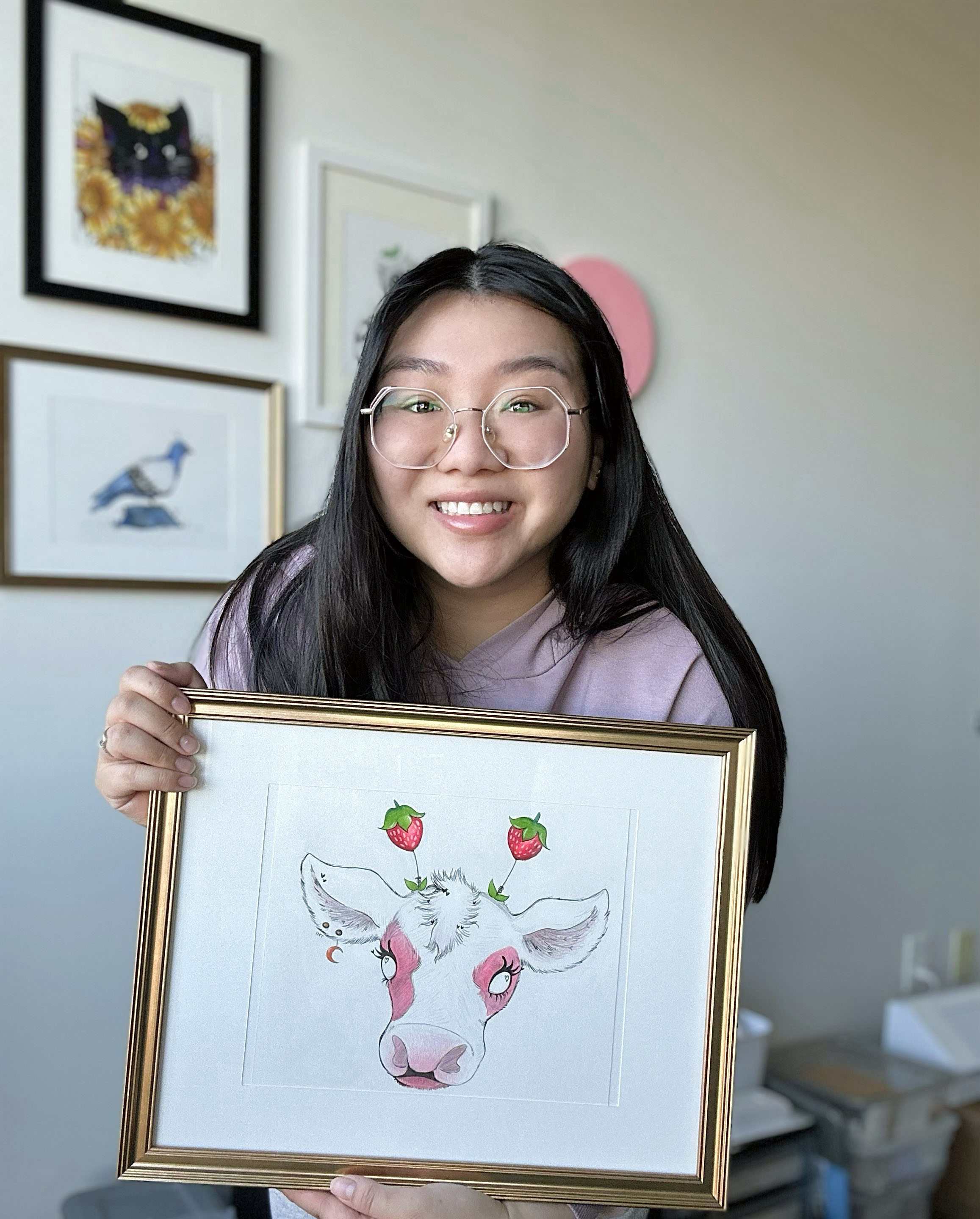 A young woman with long black hair, glasses, and a purple hoodie smiling and holding a framed artwork of a cow with pink face paint, strawberries on its horns, and a gold border. In the background, there are three framed pictures on a white wall.