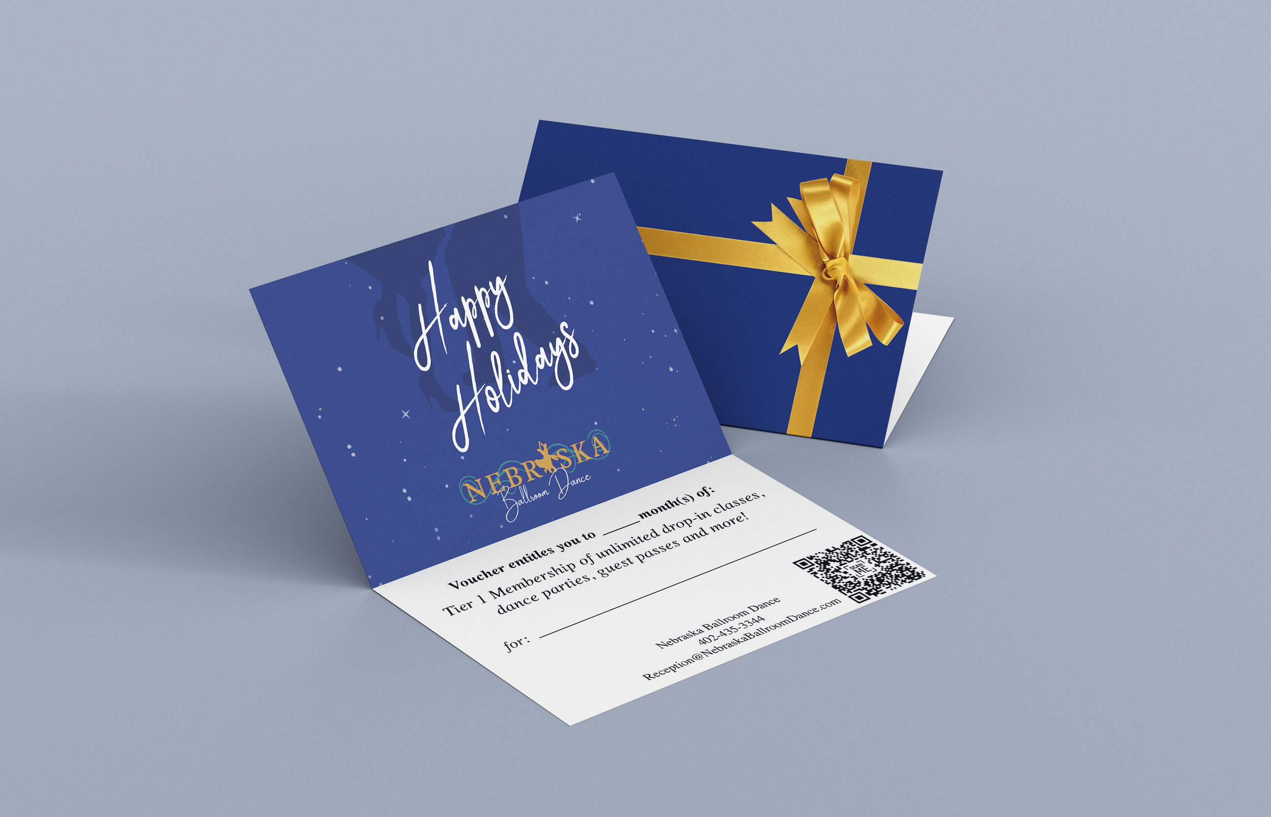A birthday or holiday gift card with a blue background, white snowflakes, and the message 'Happy Holidays' in white lettering. The card also features a logo for Nebraska Ballroom Dance and a QR code. Behind the card is a closed navy blue gift box wit